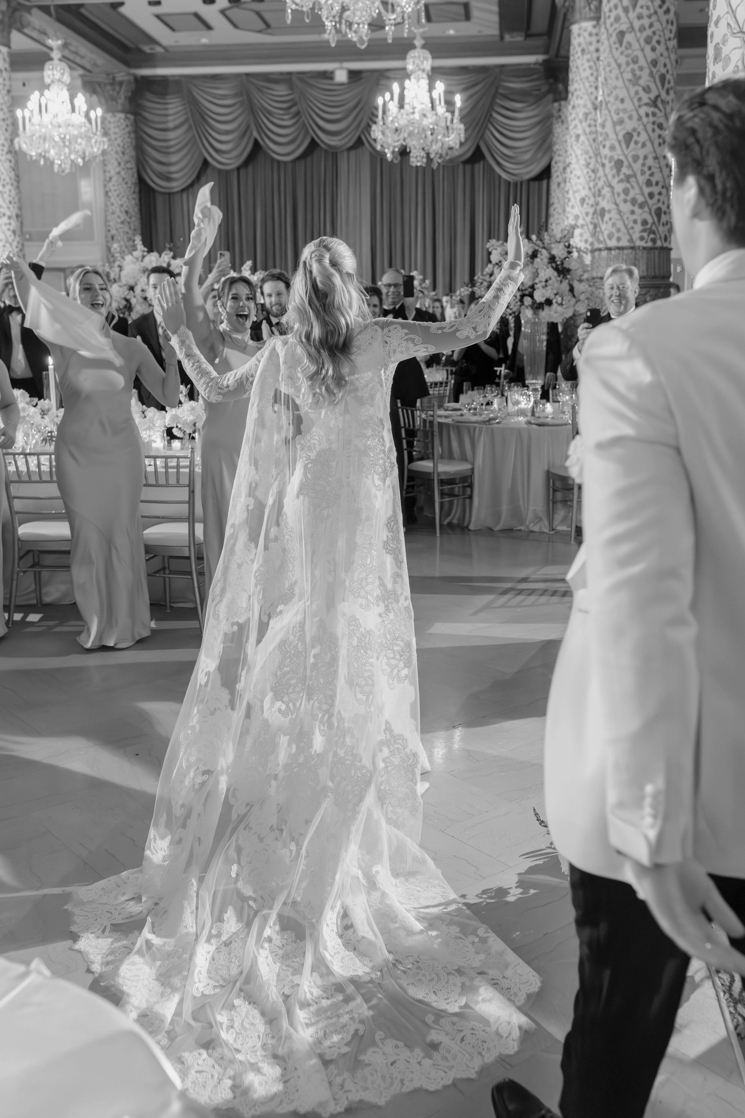 A wedding reception with a bride in a long lace wedding gown dancing with guests, surrounded by elegantly dressed people, chandeliers, and floral arrangements in a decorated ballroom.
