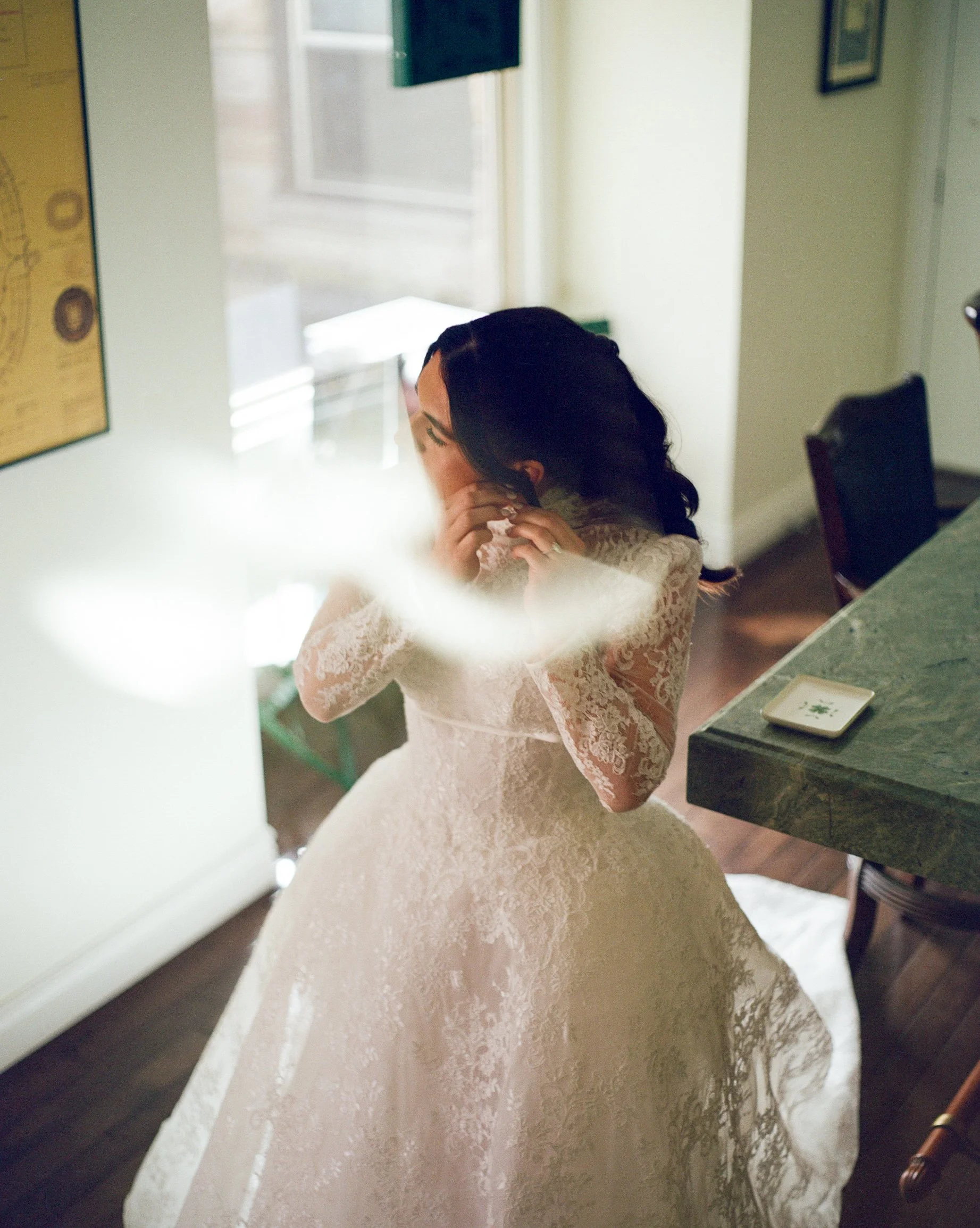 A woman in a lace wedding dress is adjusting her jewelry in a room with a mirror and a green marble countertop.