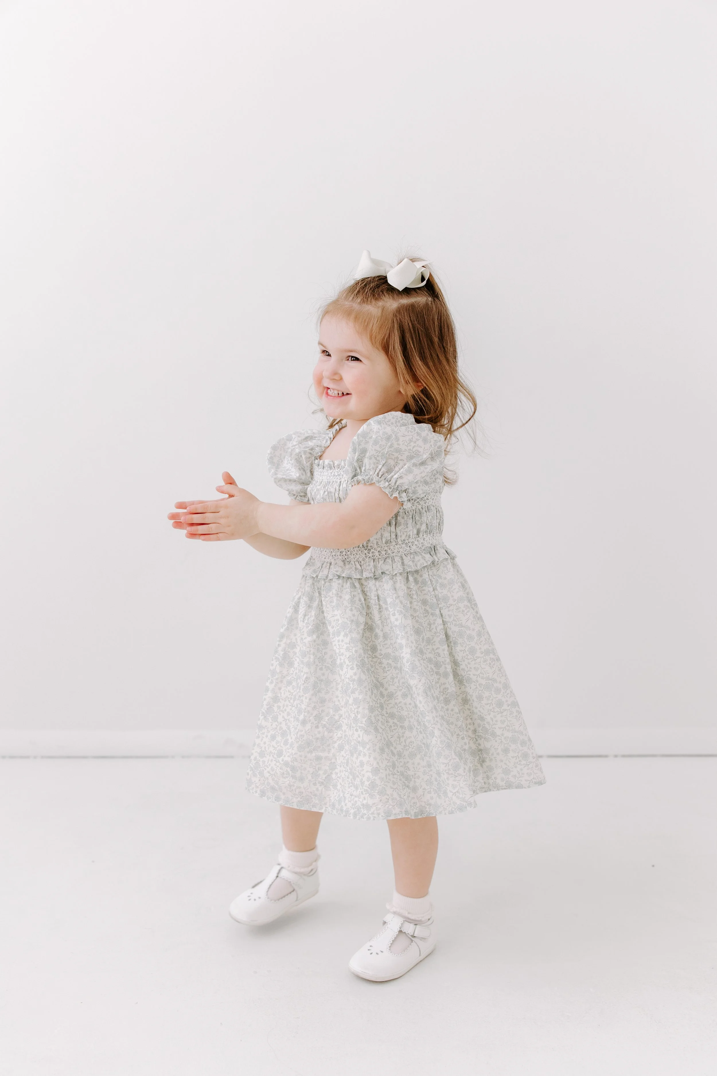 A young girl with red hair, wearing a white dress with a floral pattern, white sneakers, and a large white bow in her hair, stands against a plain white background smiling and clapping her hands.