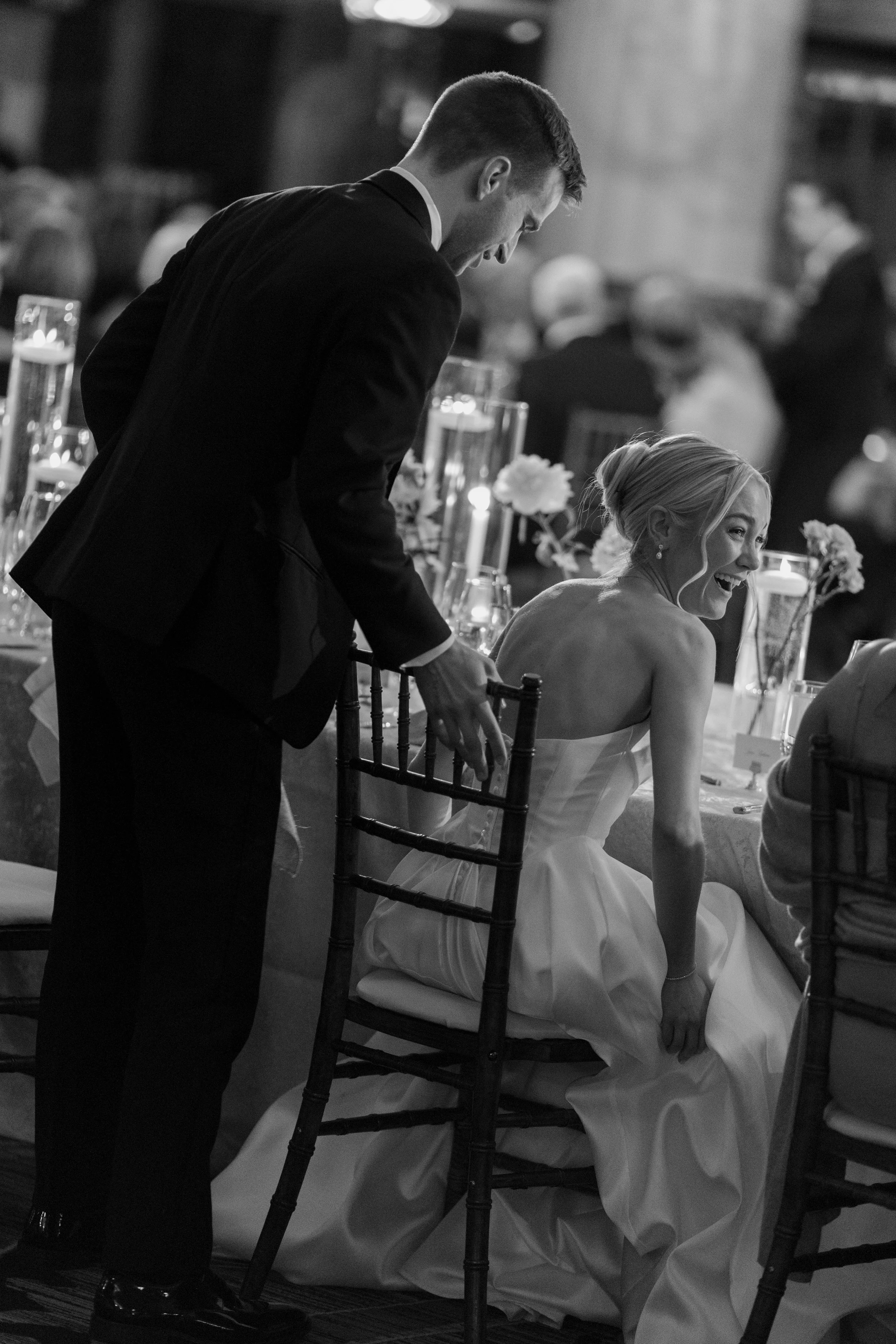 A black and white photo of a man and a woman at a formal event. The man is standing and leaning over, while the woman is seated wearing an elegant dress and laughing. The table is decorated with candles and flowers.