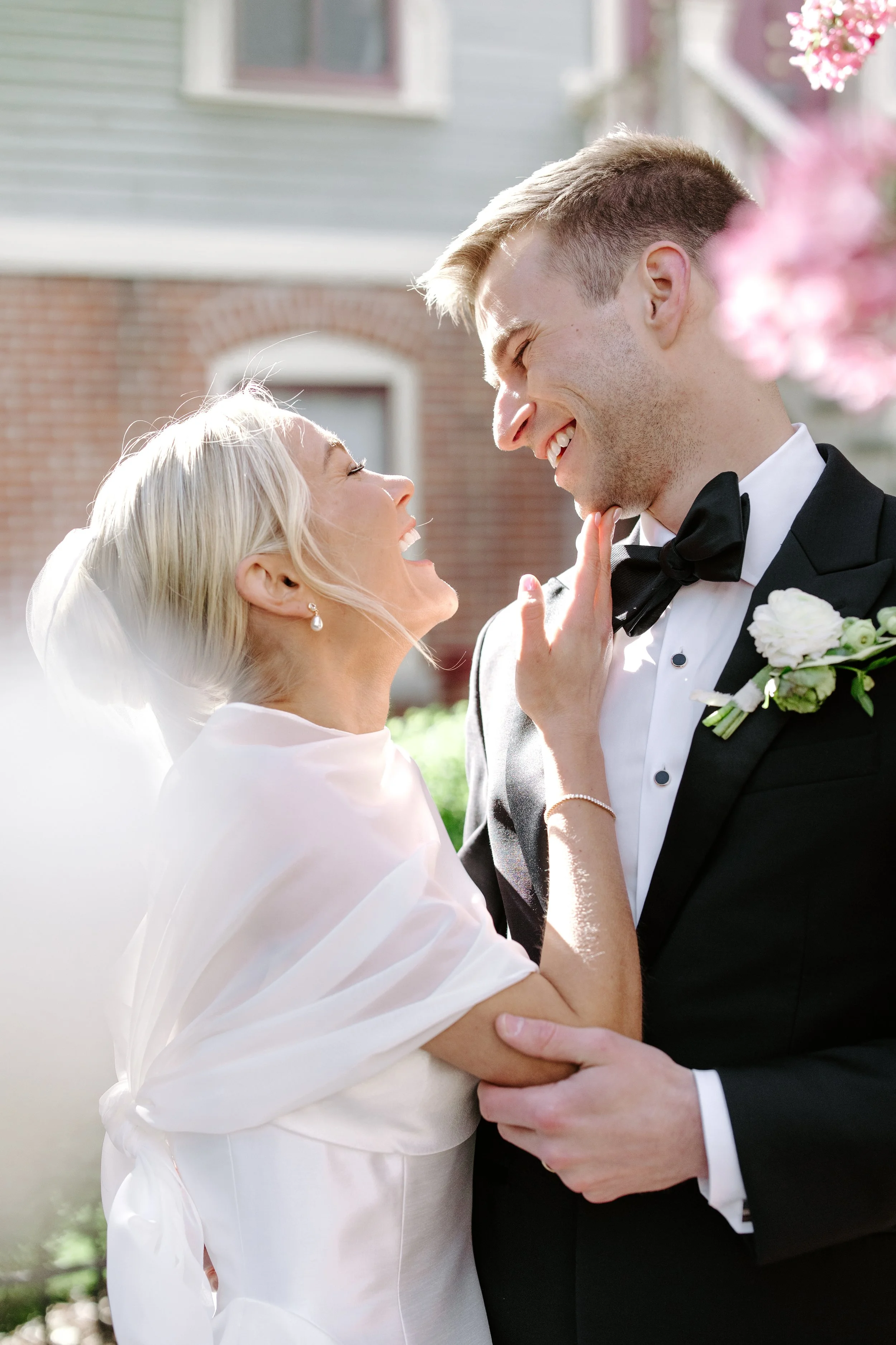 A happy couple is embracing outdoors on a sunny day, with the woman wearing a white dress and the man in a black tuxedo with a bow tie. The man is smiling and touching the woman's chin, while she has her eyes closed and is smiling back.