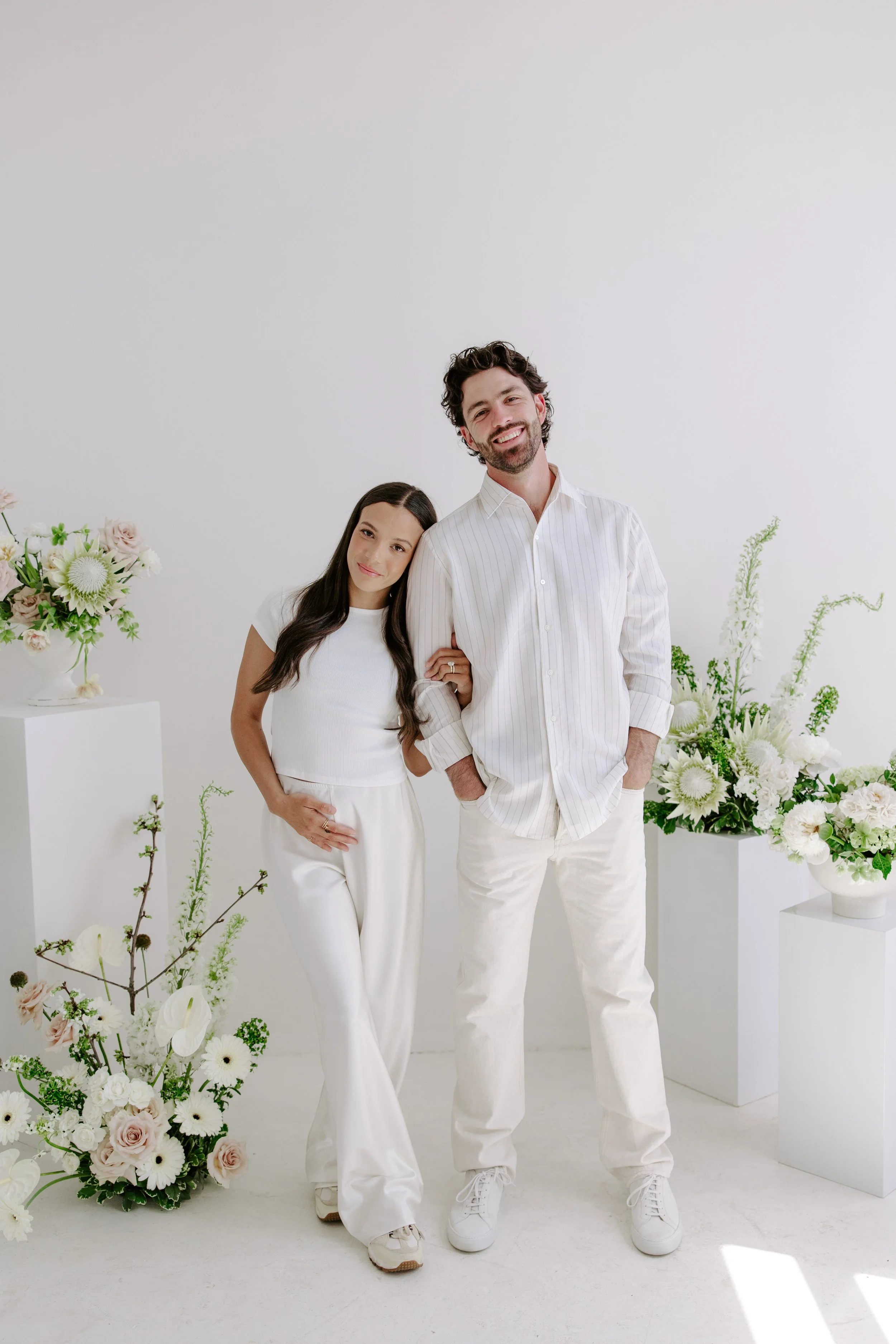 A smiling man with curly dark hair and beard, wearing a white striped shirt and white pants, standing next to a woman with long dark hair, wearing a white top and white pants, in a minimalist white room with various white and pastel-colored floral ar