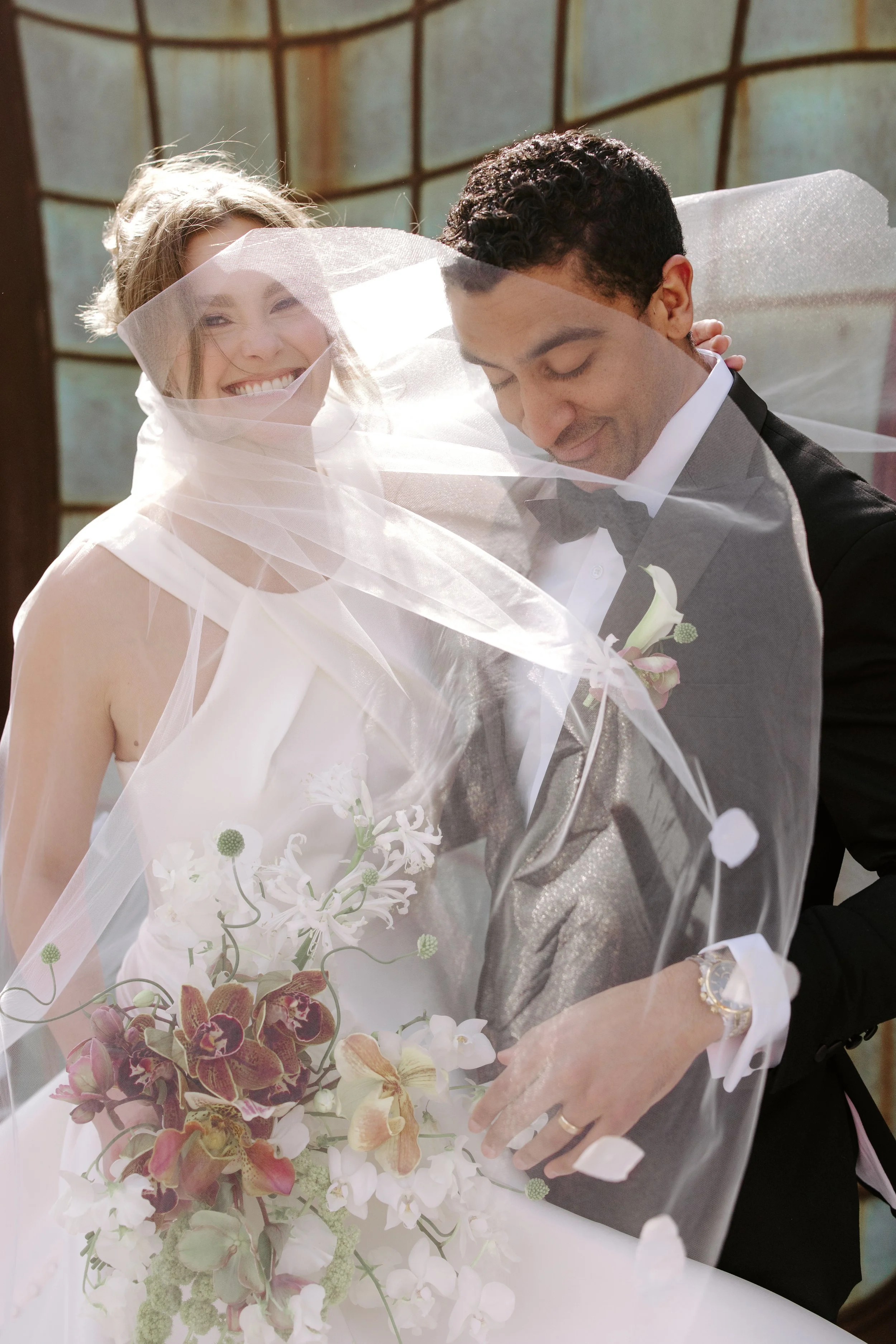 A bride and groom sharing a joyful moment under her veil. The bride is smiling widely and holding a bouquet of flowers, while the groom has his eyes closed, wearing a black tuxedo with a bow tie, and a boutonniere. They are in a bright setting with a