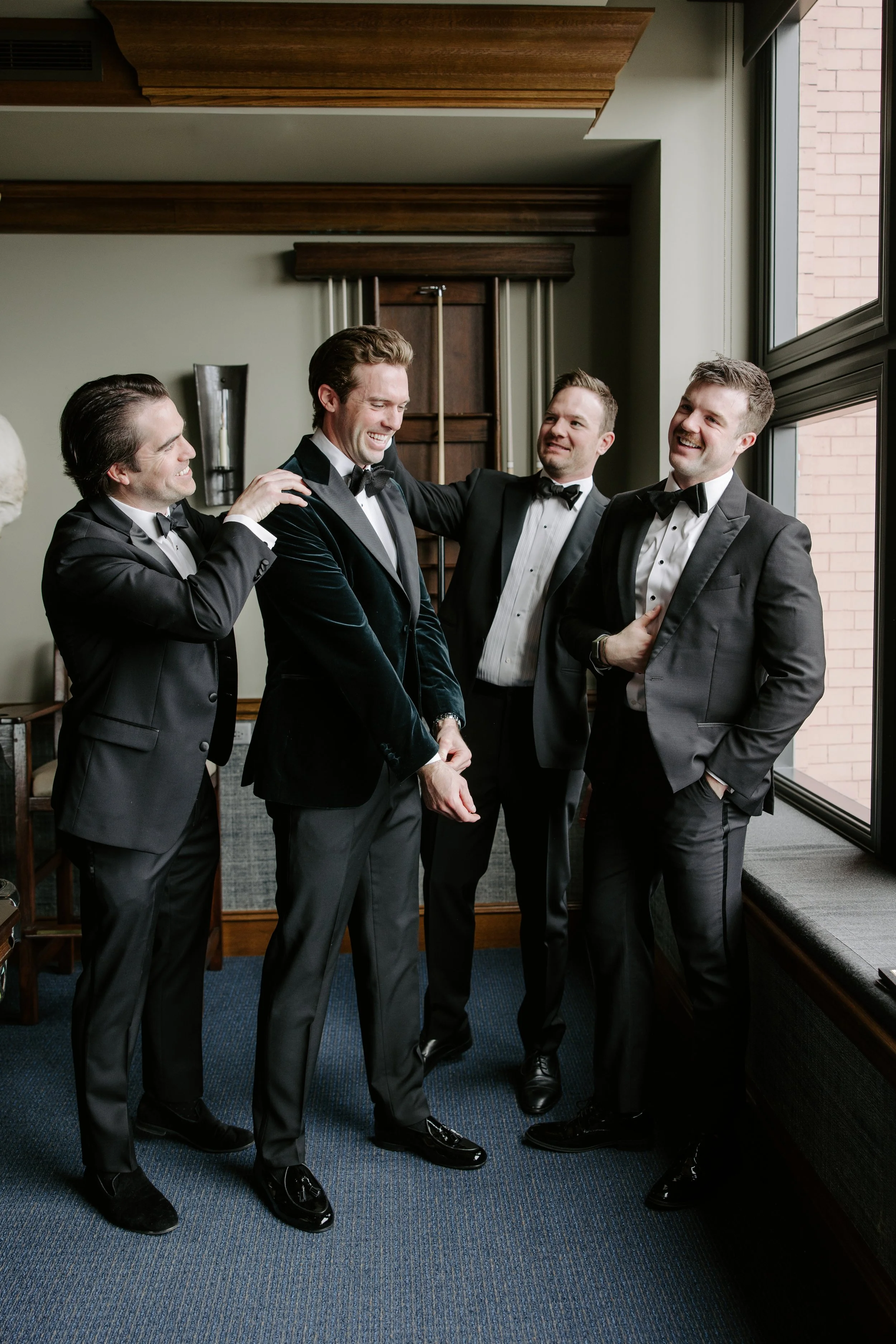 Four men dressed in tuxedos are gathered by a window. They are smiling and appear to be enjoying a moment of camaraderie.