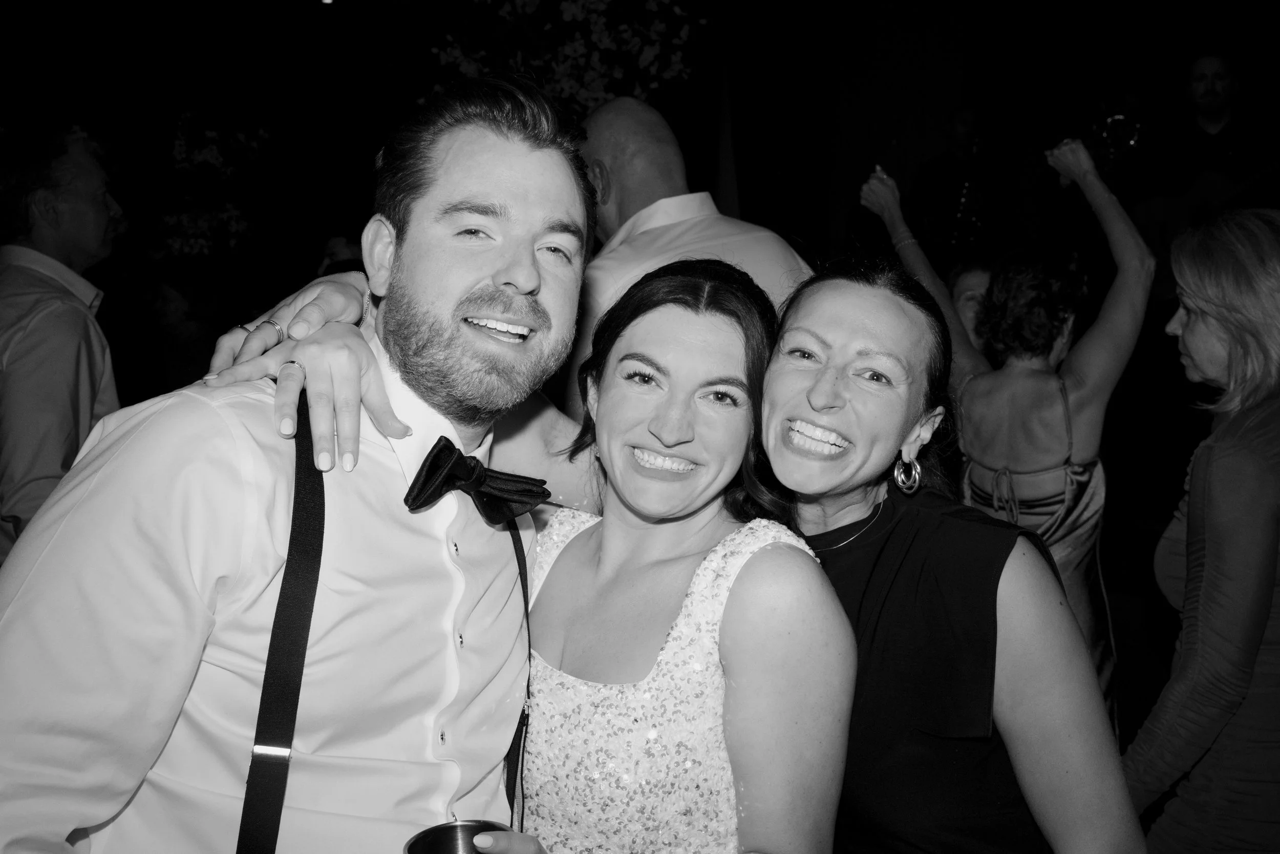 A group of three smiling people posing at a party, with a man wearing a tuxedo and two women in dresses, standing close together in a crowded social event.