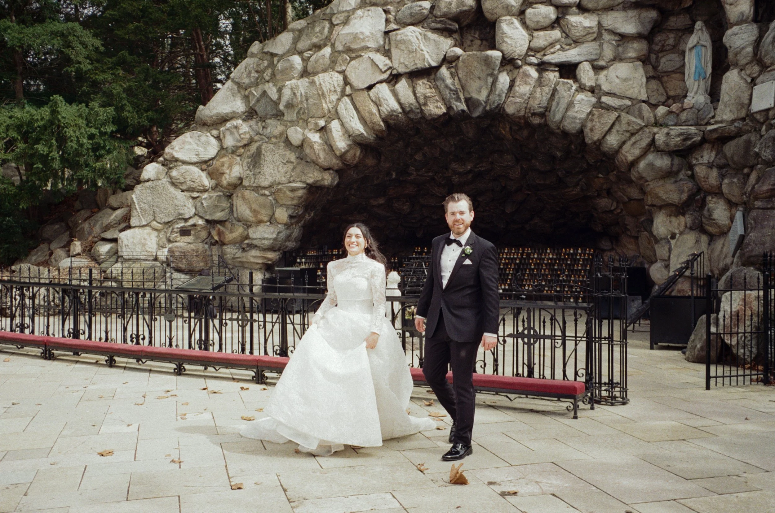A newlywed bride in a white wedding gown and groom in a black tuxedo standing outdoors in front of a stone grotto, smiling.