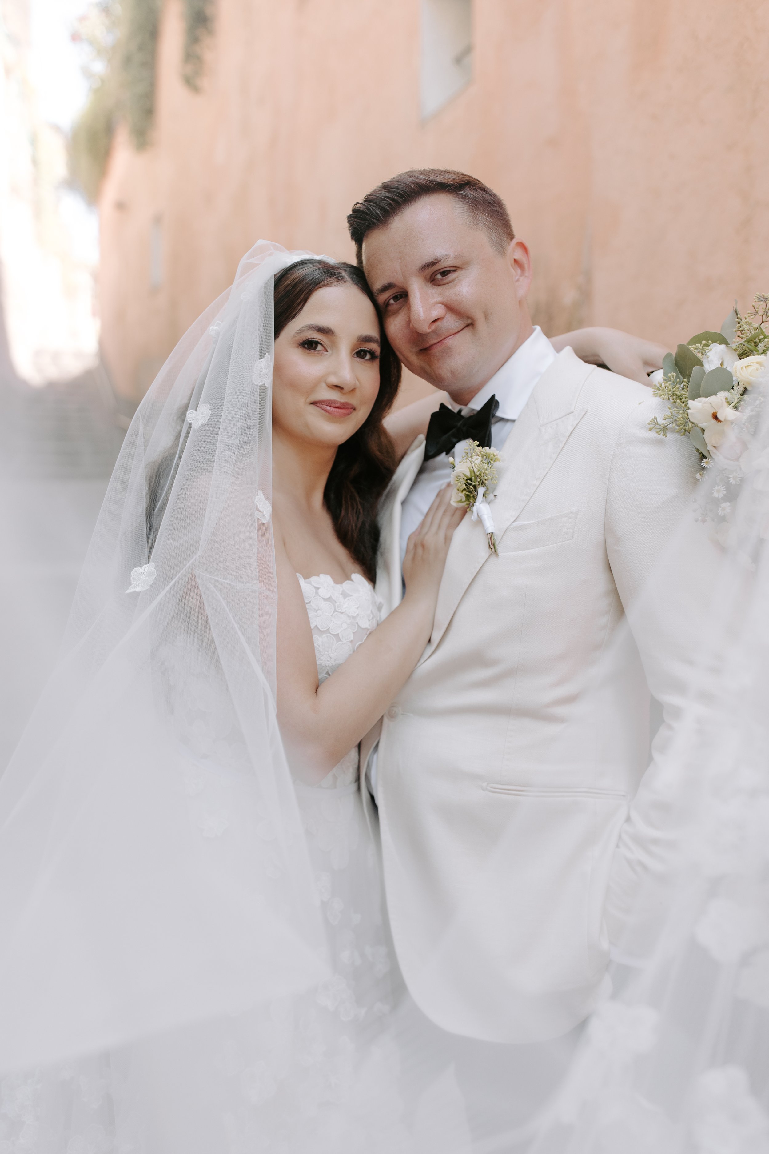 A bride and groom in wedding attire, smiling and close together, with the bride wearing a veil and the groom in a white tuxedo with a black bow tie, standing against a neutral background.