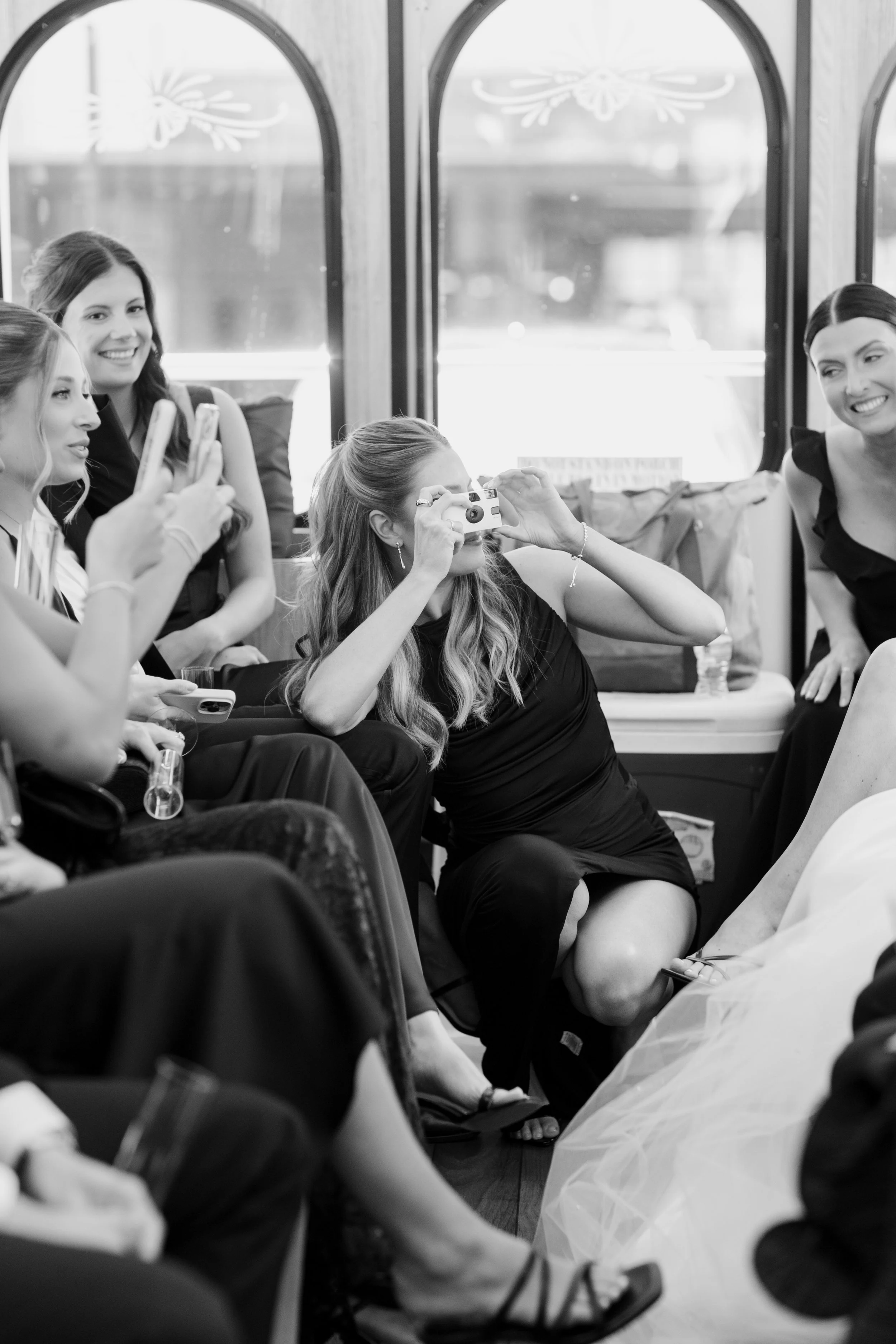 Women in elegant dresses celebrating at a formal event, with one woman taking a picture with a vintage camera, sitting on a decorated bus or trolley with large windows and ornate designs.