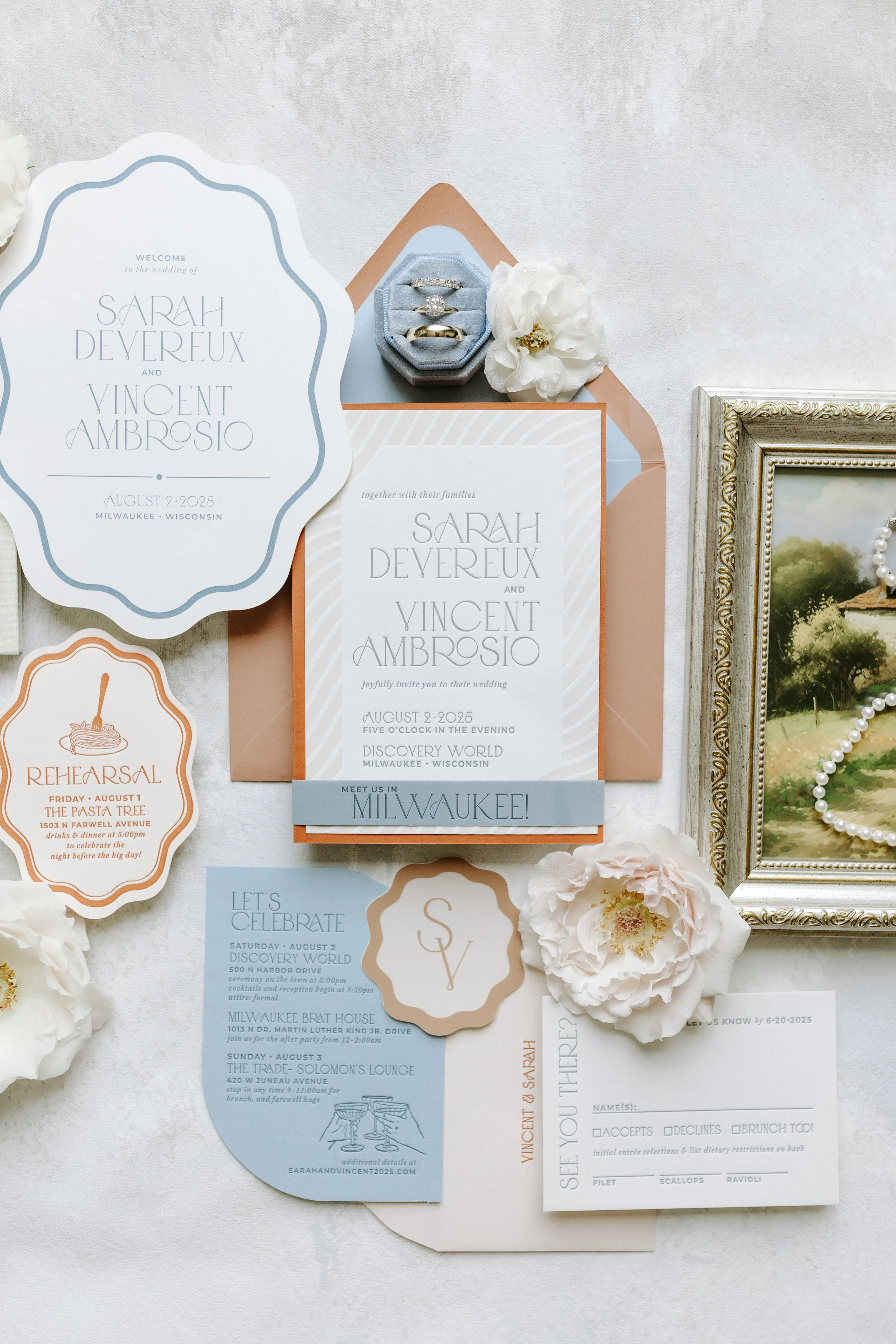 Wedding invitation suite including an announcement card, RSVP card, and additional info cards, with rings, a flower, and a framed photograph nearby, all arranged on a white background.