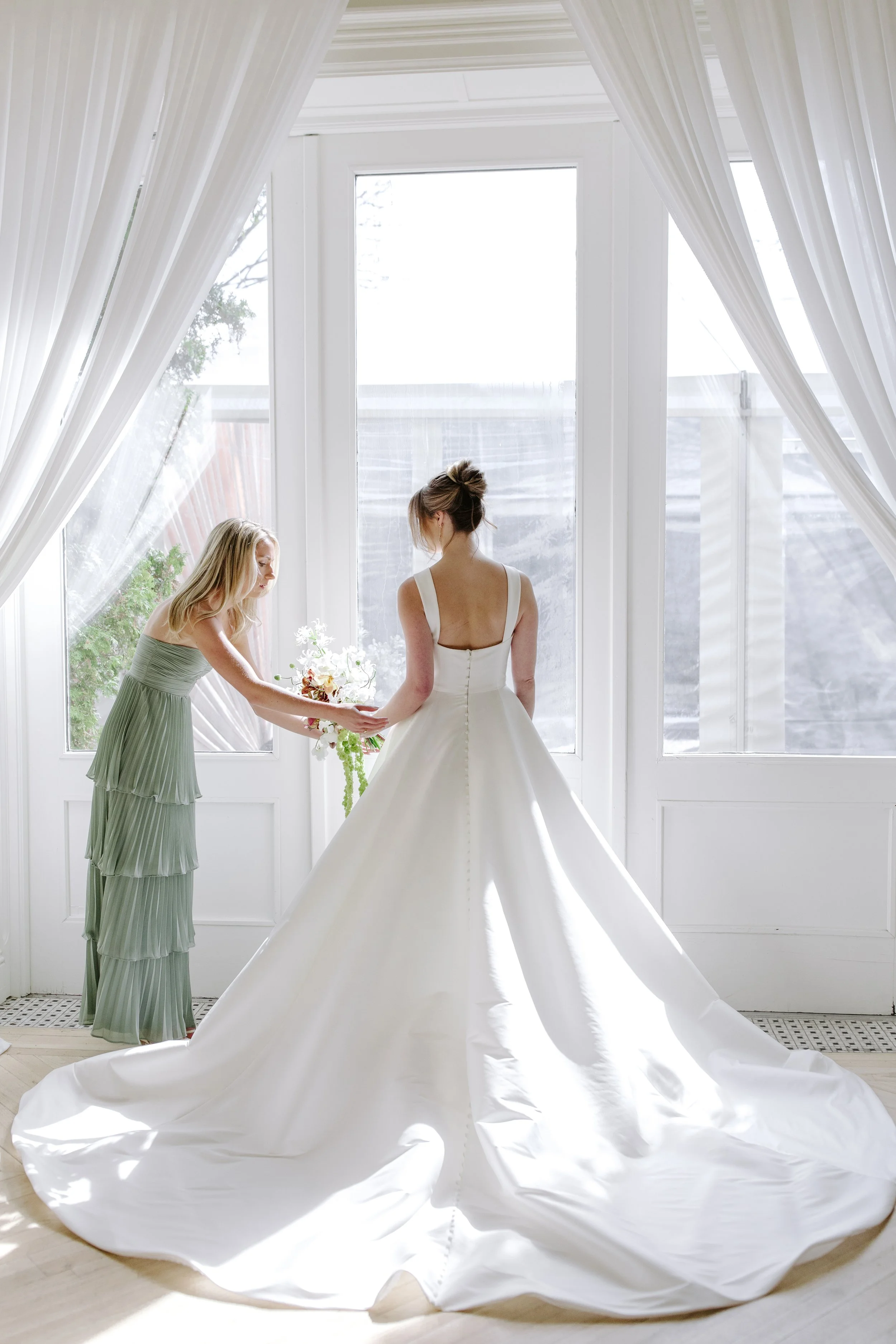 Two women in a bright, white room with large windows, one in a wedding dress and the other in green, holding the train of the bride's dress and a bouquet or floral piece.