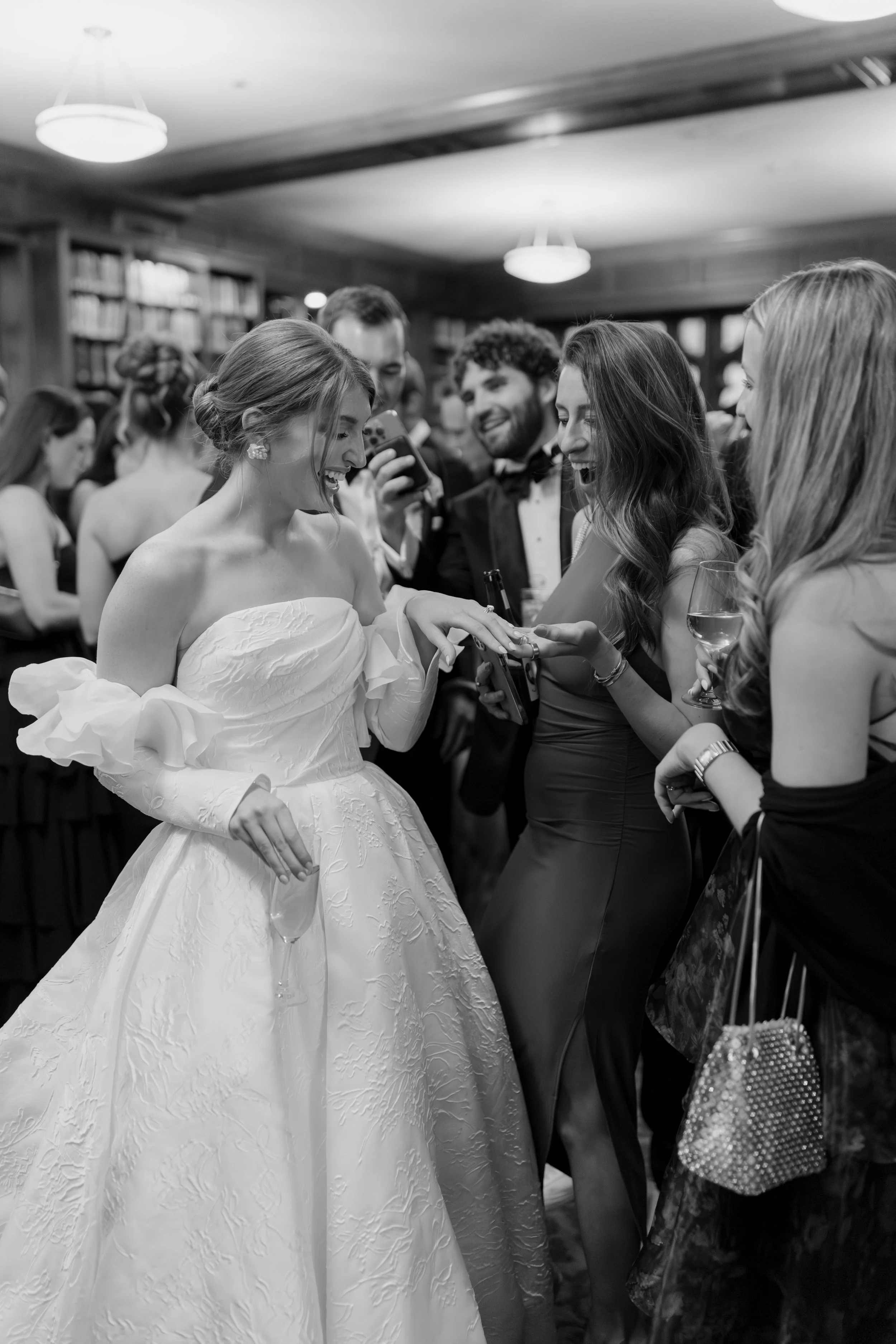 A bride in a wedding dress is smiling and holding a glass of wine as she interacts with guests at her wedding reception, surrounded by friends dressed in formal attire, with a background of bookshelves and light fixtures in a cheerful atmosphere.