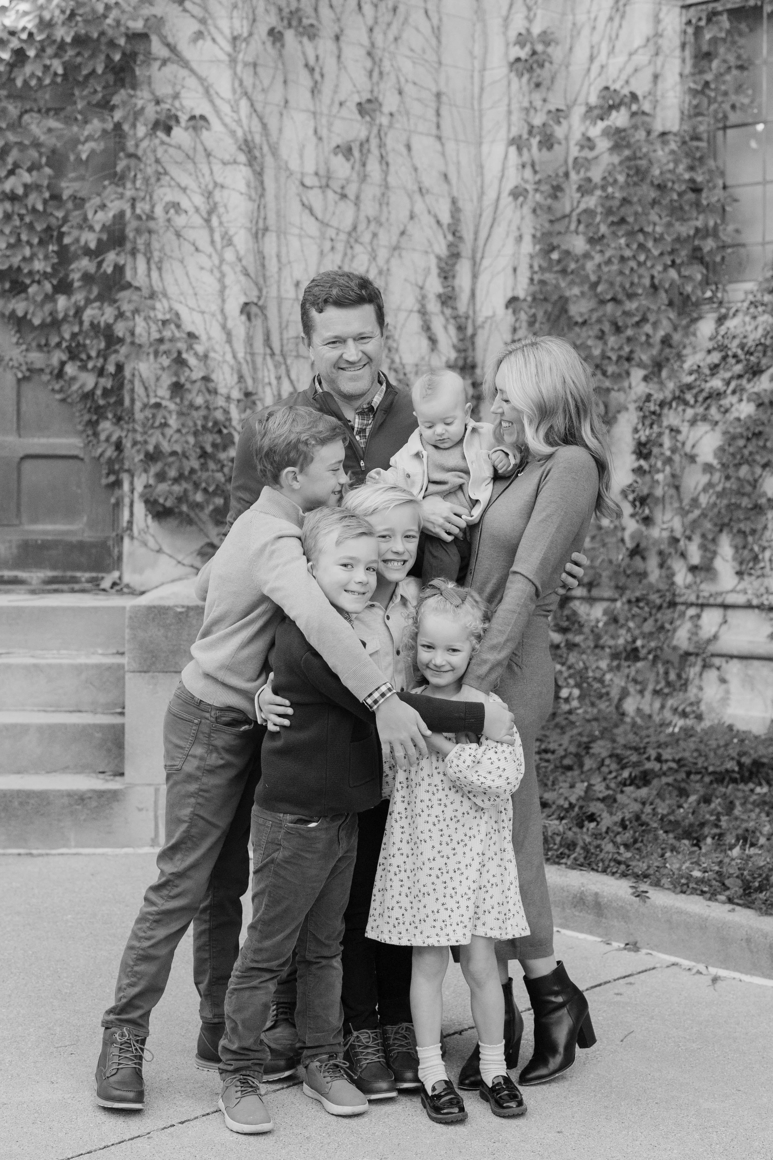 A family of eight posing outdoors on a sidewalk in front of leafless trees and a brick wall covered with vines. The group includes three adult women and five children, smiling and hugging each other.