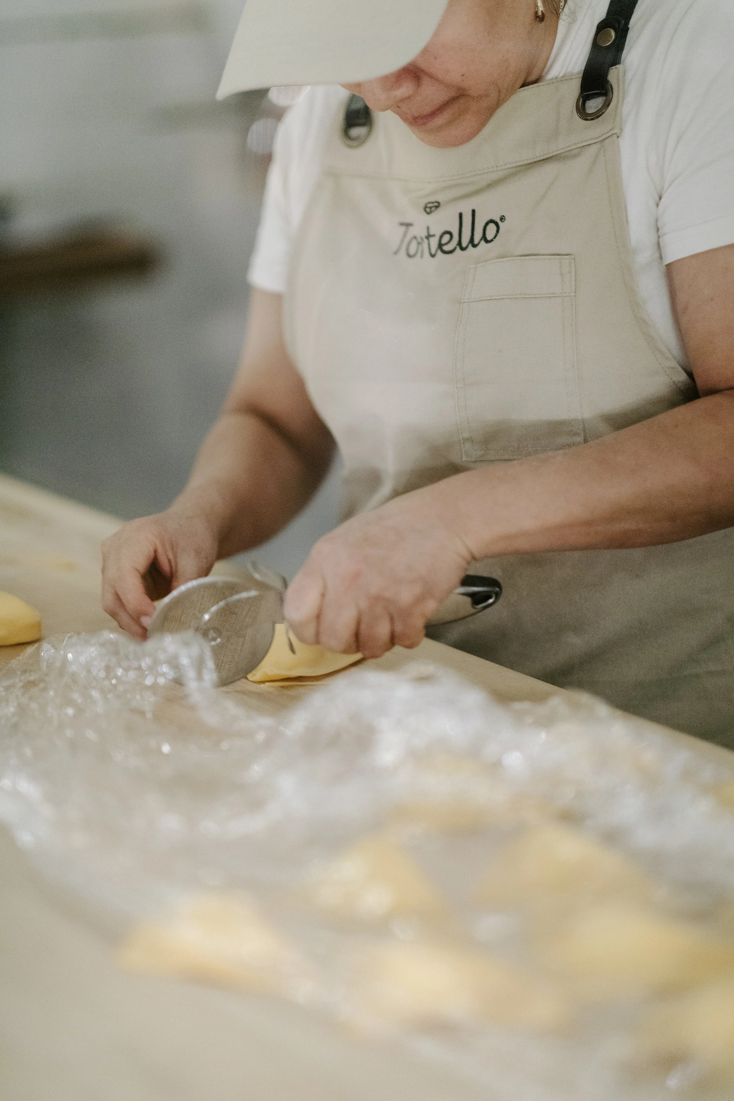 A woman wearing a white toque and apron labeled 'Tortello' is preparing pasta dough on a marble countertop. She is using a round cutter to cut out pasta circles, with flour and dough visible on the surface.