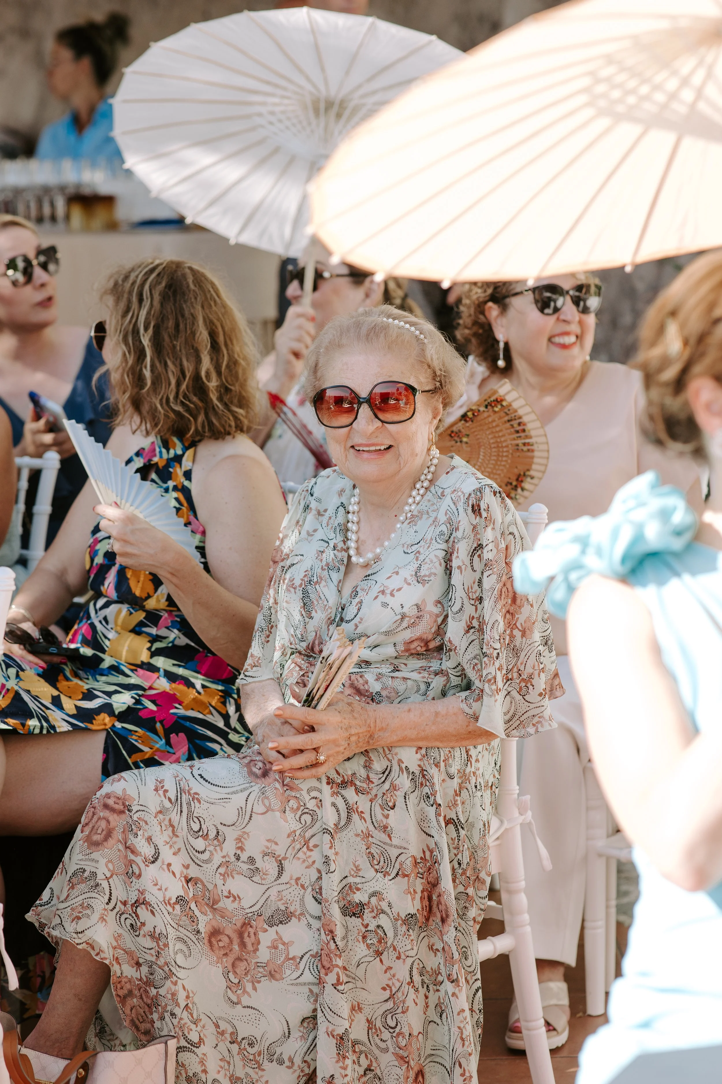 Group of women sitting outdoors at a social event, some holding parasols, wearing sunglasses, and dressed in light, floral, and elegant clothing.