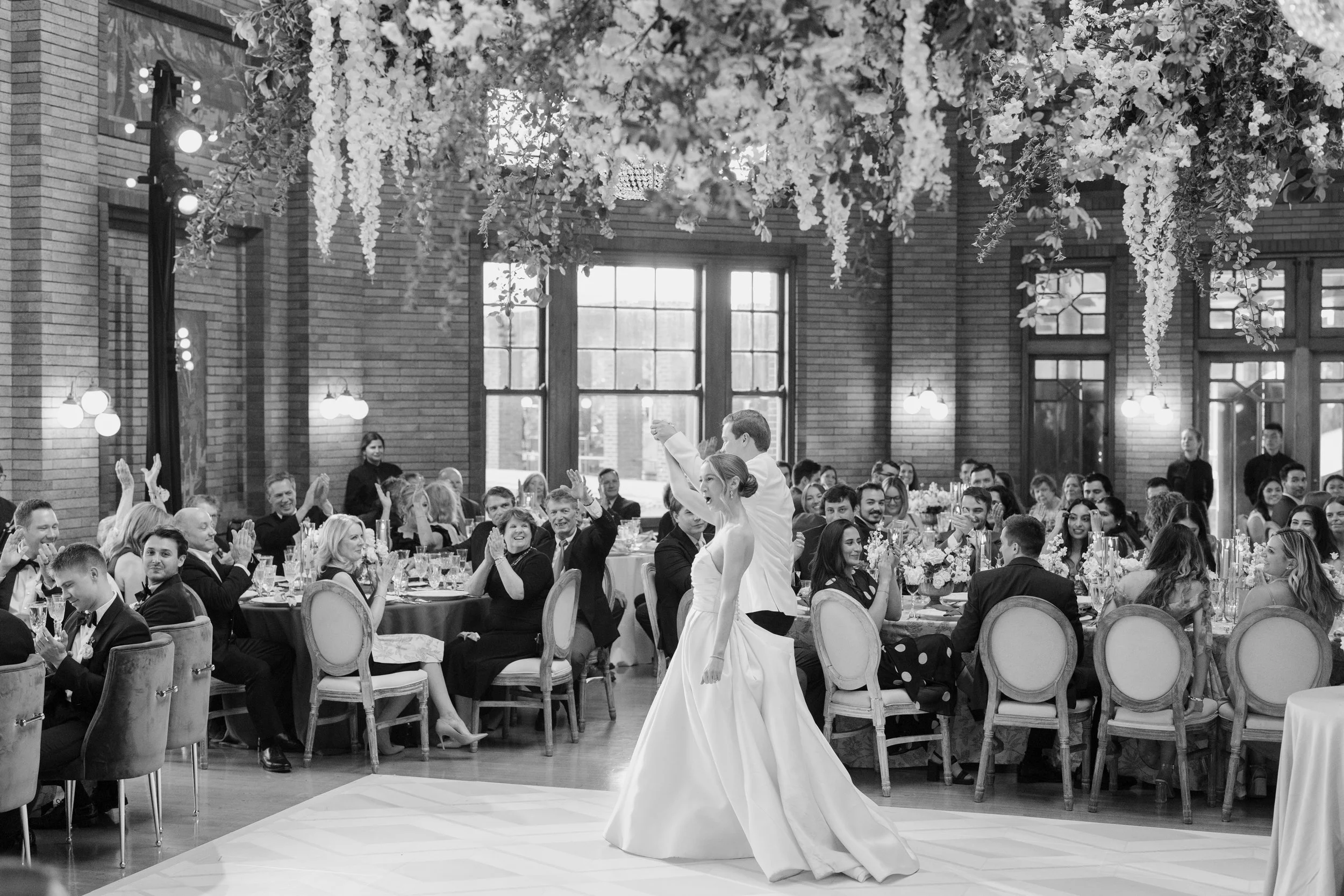 A wedding reception with a bride in a white gown dancing in a grand hall with high windows and brick walls, surrounded by seated guests at decorated tables.