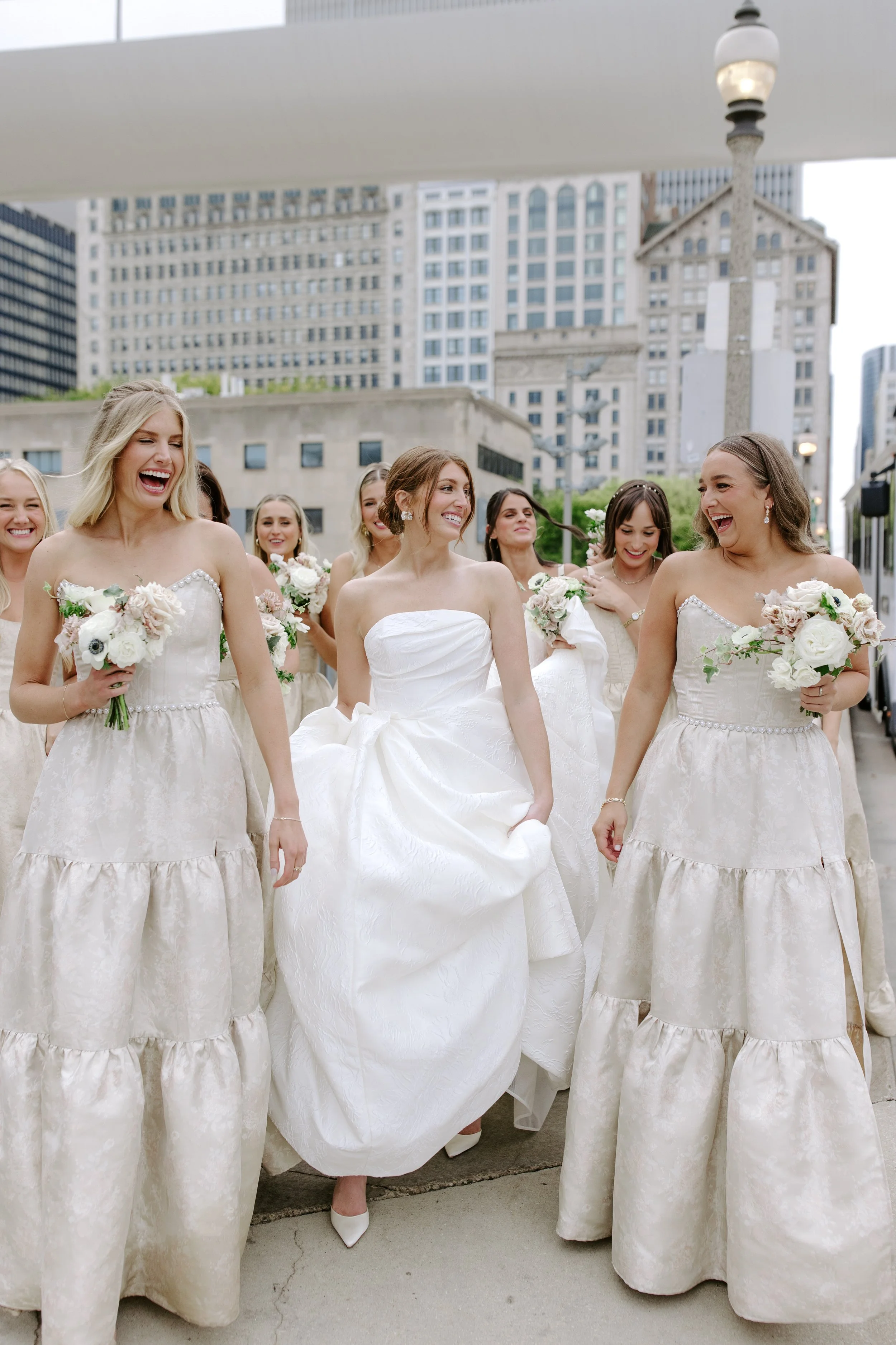 Group of women in wedding dresses celebrating outdoors in a city, holding bouquets, with tall buildings and a lamp post in the background.