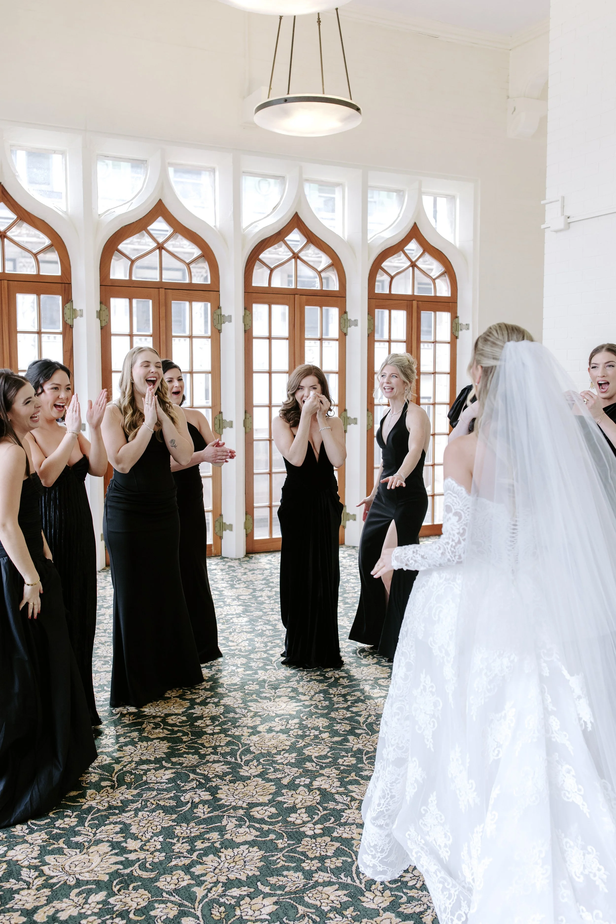 A bride in a white lace wedding gown and veil stands facing a group of women in black dresses who are amused and smiling.