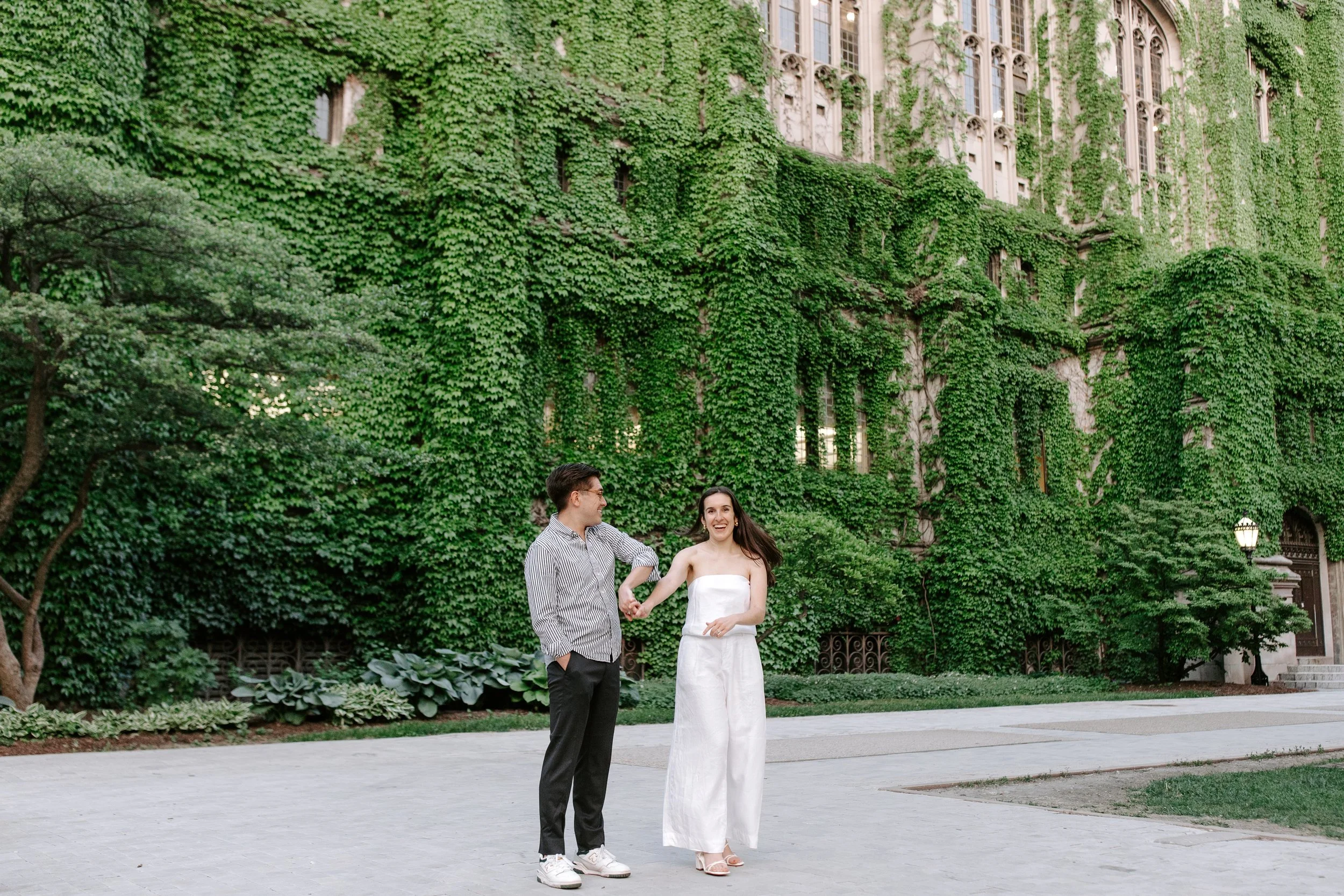 A couple is standing outside in front of a building covered with green ivy, holding hands, smiling, dressed in casual attire.