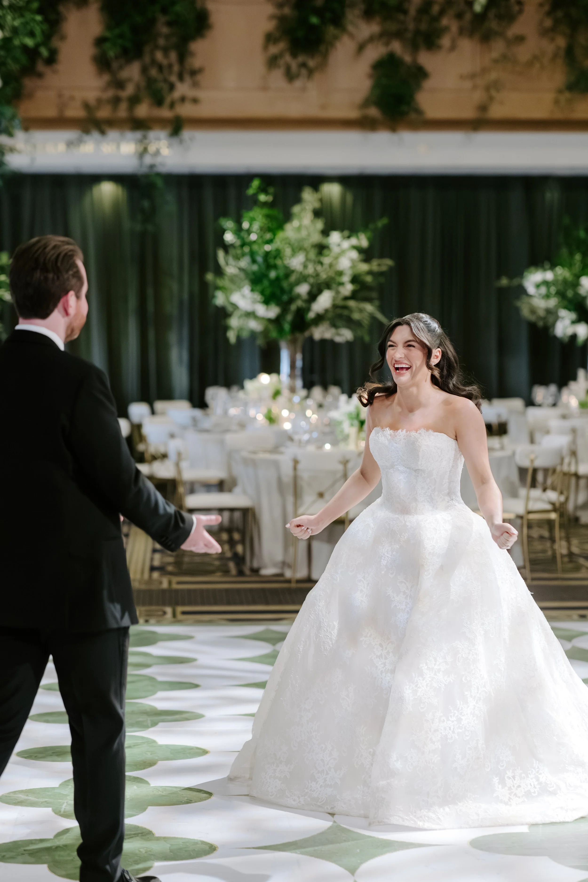 A bride in a white wedding gown smiling and dancing with a groom in a black suit during a wedding reception in a decorated banquet hall.