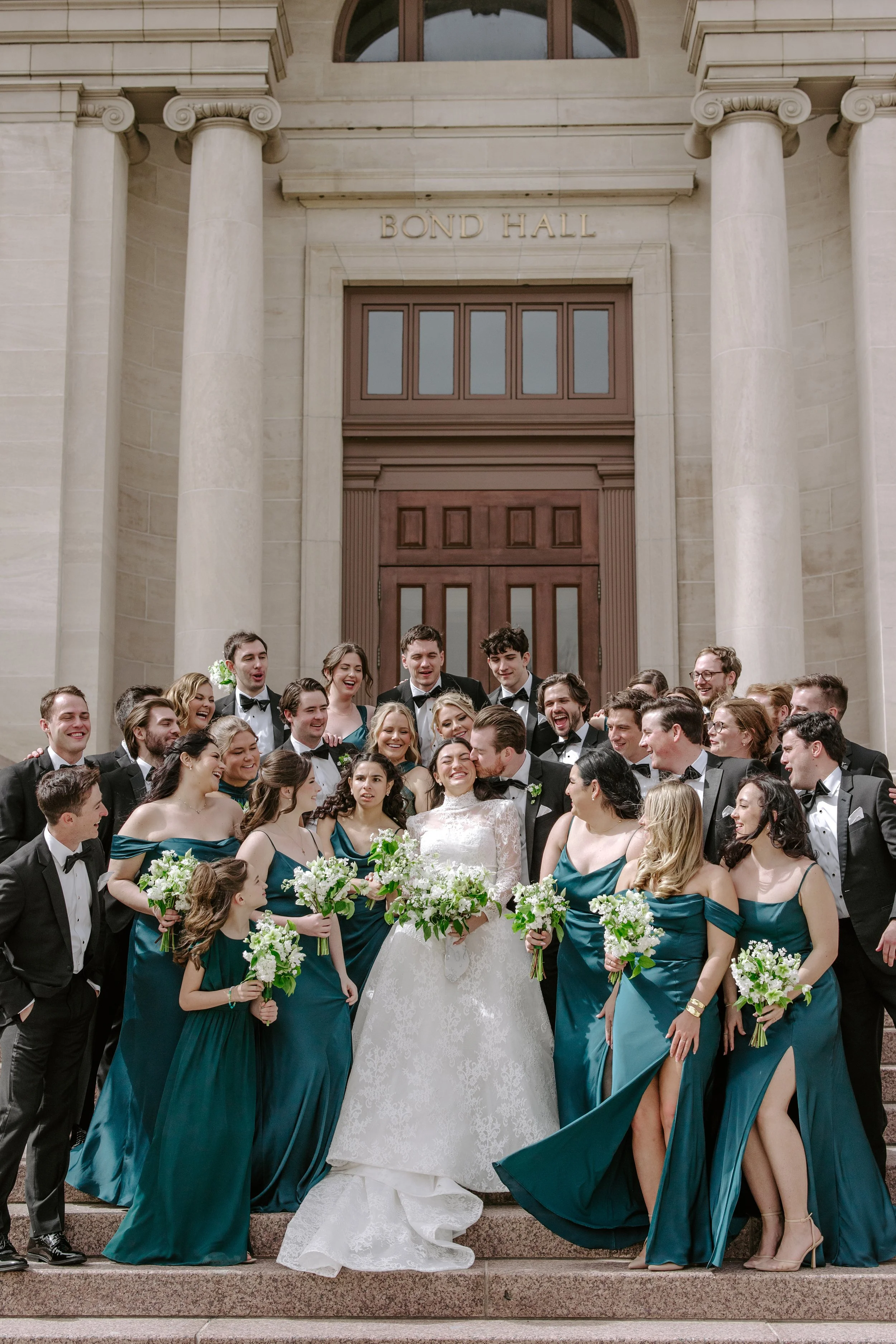 A wedding group photo on the steps of a building with the sign 'Bond Hall' above the door. The group includes the bride and groom surrounded by bridesmaids and groomsmen, all dressed in formal attire. The bridesmaids are wearing teal dresses, and the