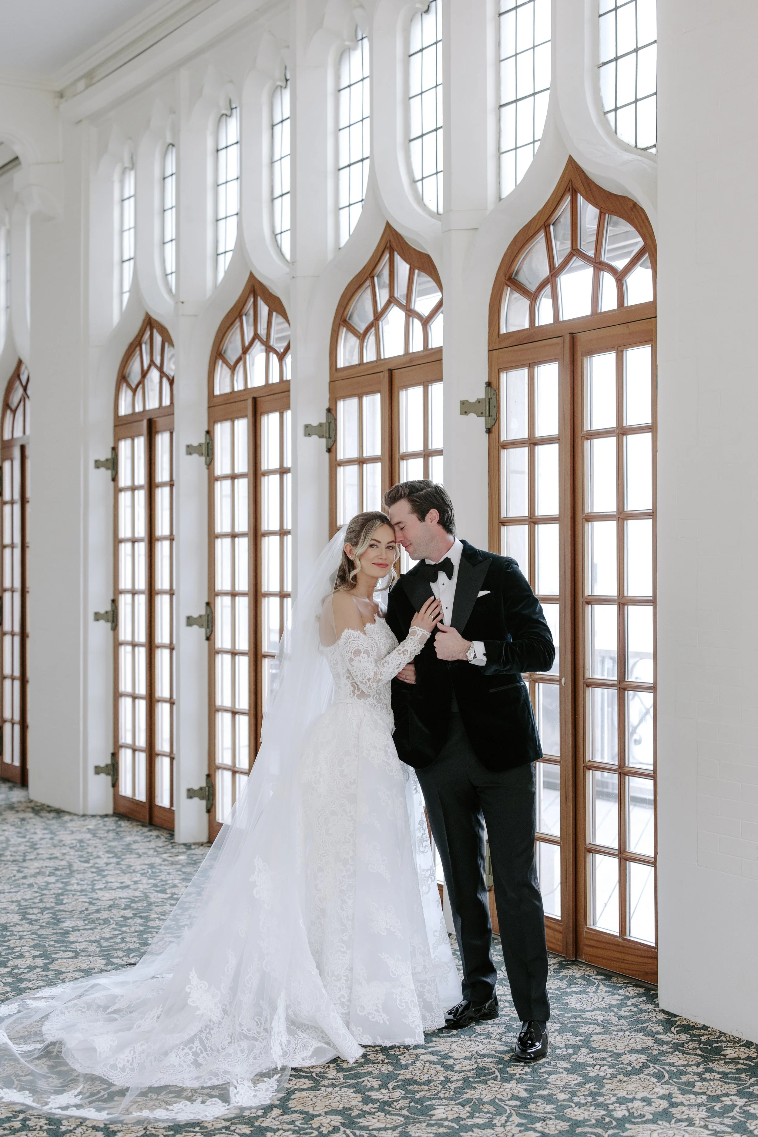 A bride and groom in wedding attire standing close together inside a bright room with large windows featuring arched and rectangular wooden frames.