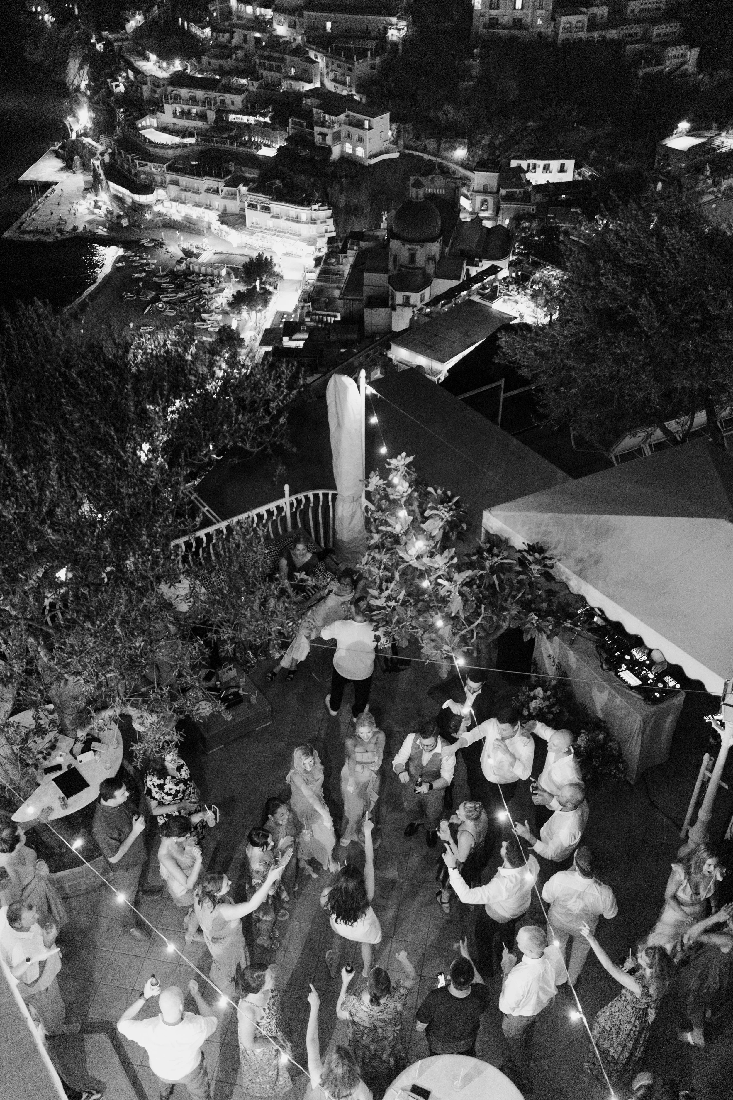 Nighttime scene of an outdoor party with people dancing and socializing on a dance floor surrounded by string lights, with a cityscape of buildings and boats visible in the background.