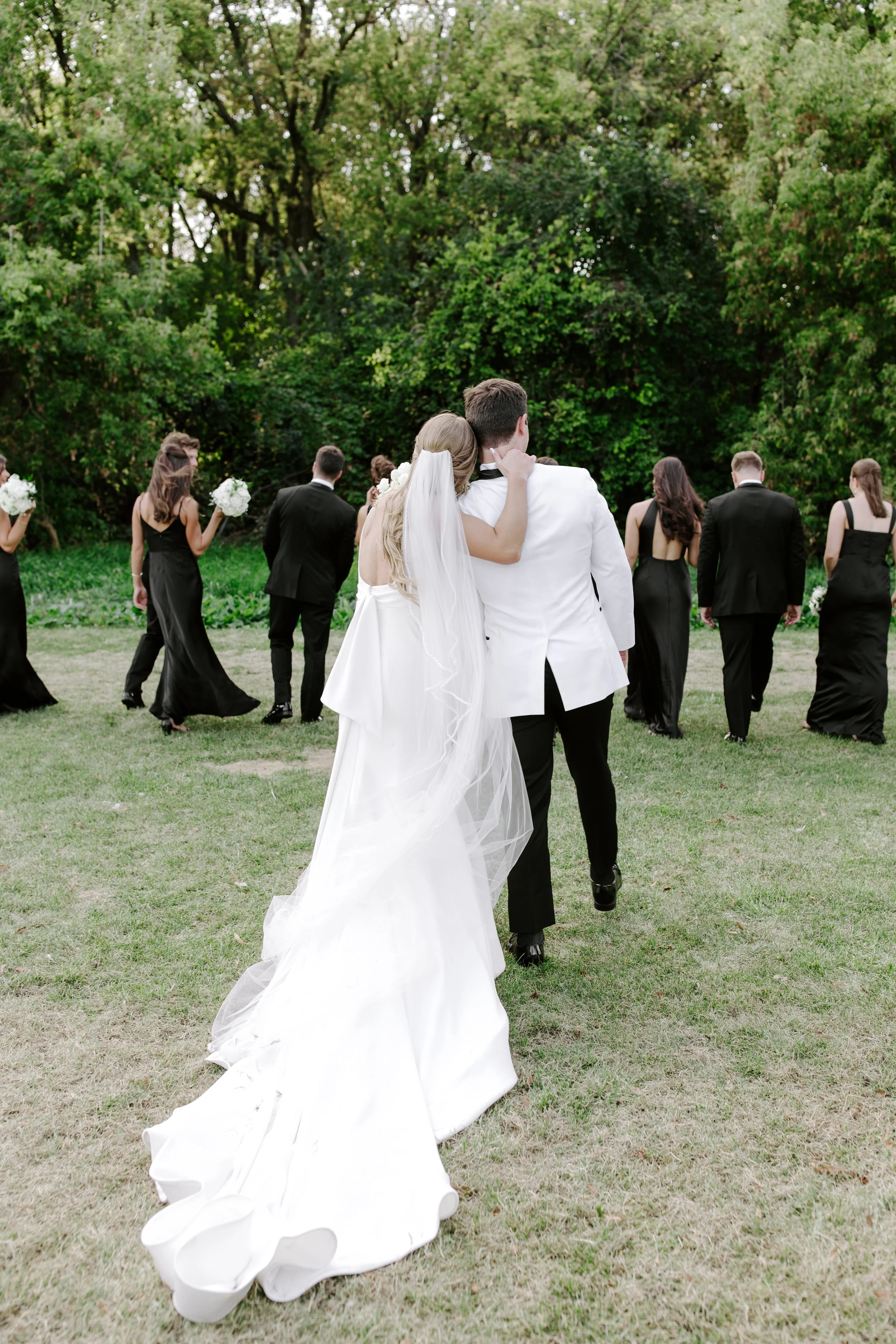 A bride and groom holding each other as they walk through a grassy area with trees, during their outdoor wedding ceremony, with bridesmaids and groomsmen in the background.