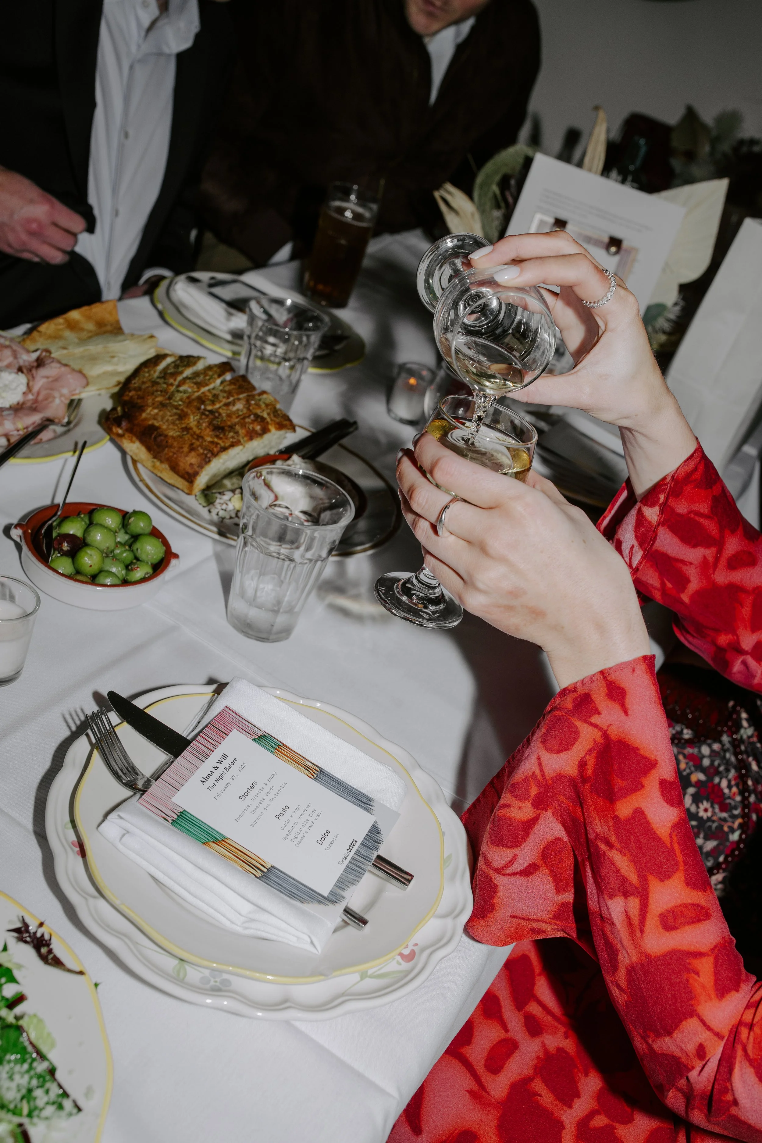 A person in a red patterned dress is pouring water from a glass into a wine glass on a dining table set for a meal. The table has plates with food, utensils, glasses, and a menu. The setting appears to be a formal dinner event.