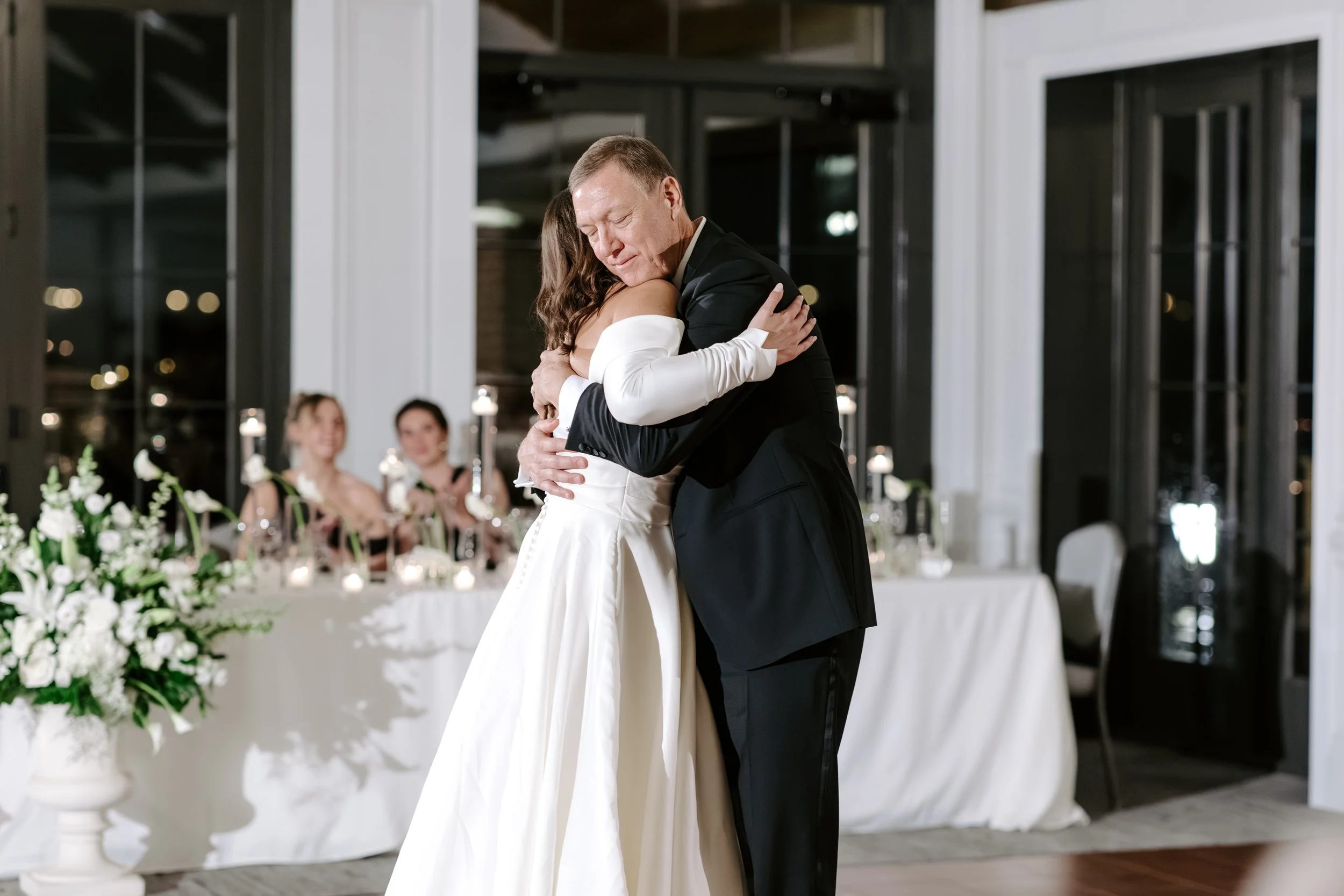A bride and groom share a dance at their wedding reception, with wedding guests seated at decorated tables in the background.