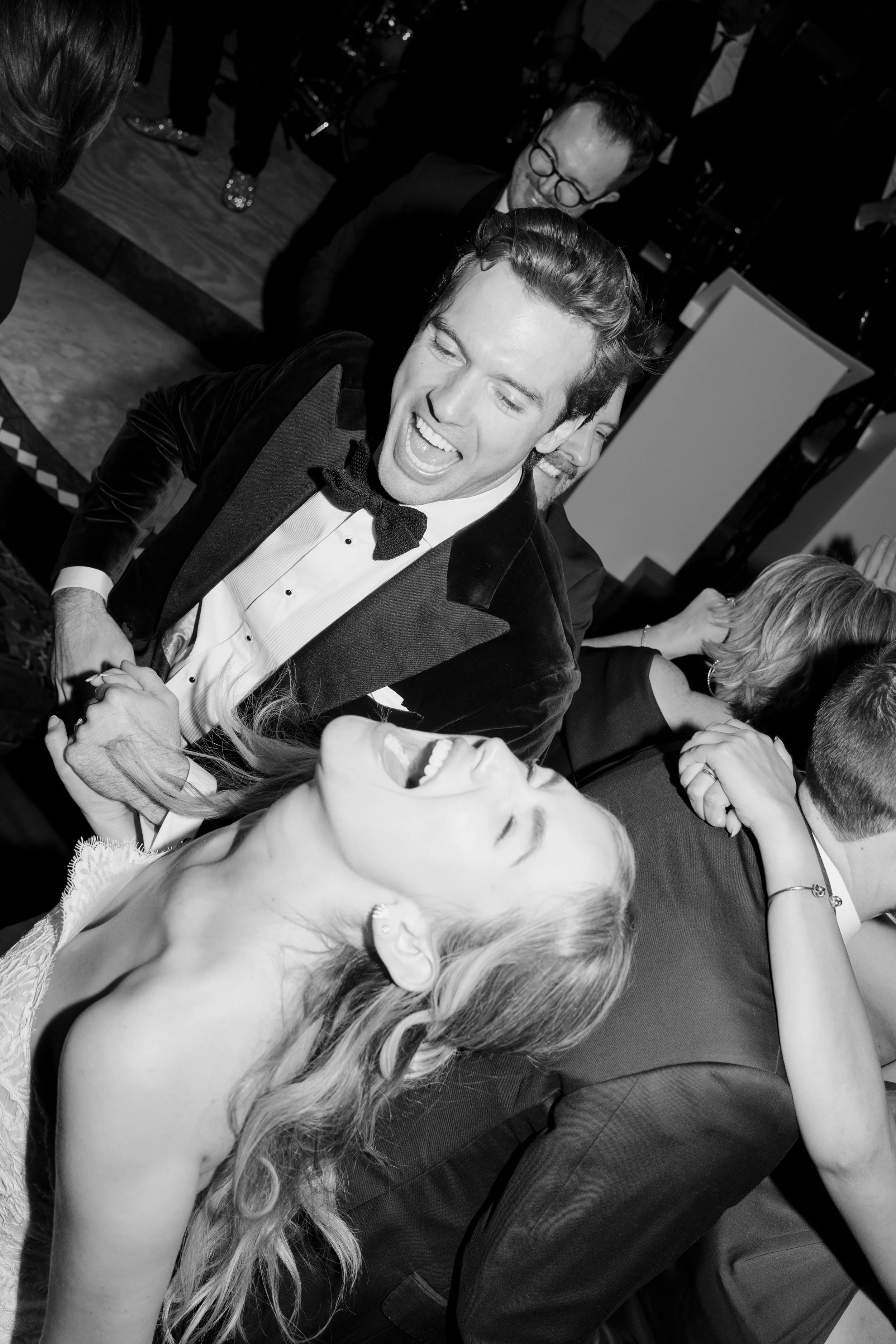 Black and white photo of a man in formal attire with a bow tie dancing and smiling with a woman with long wavy hair, dressed in an elegant gown at a festive event.