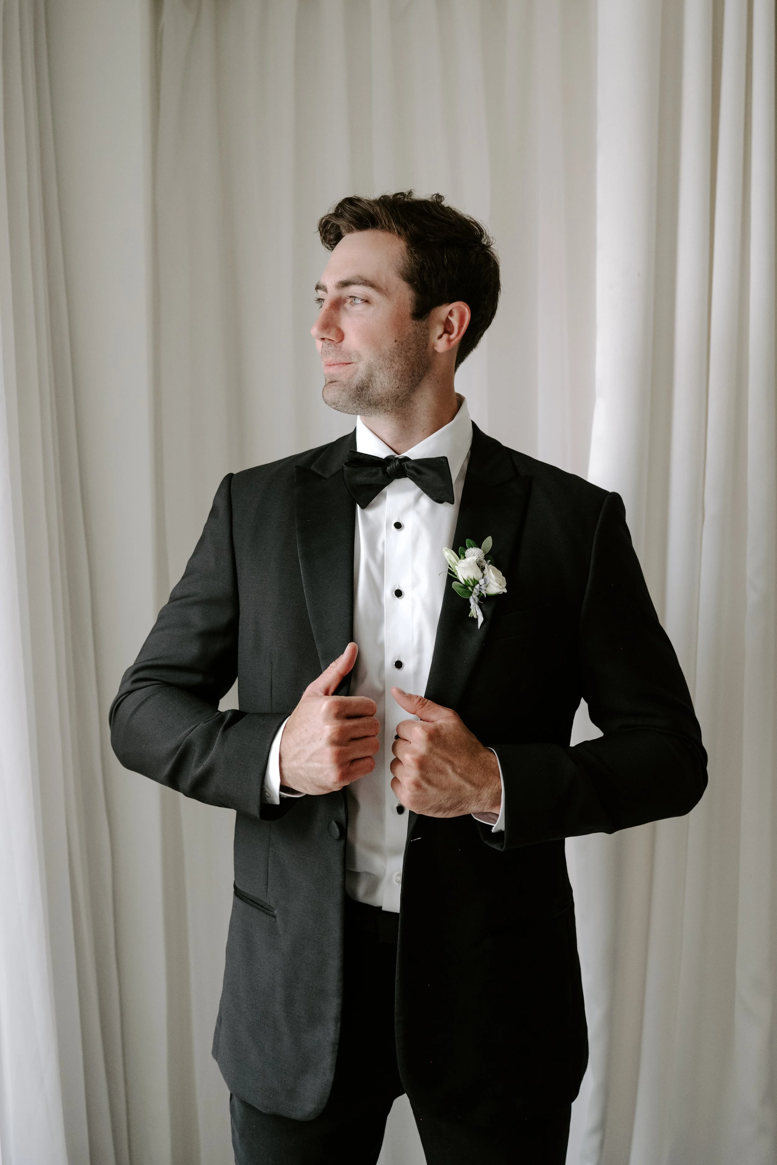 A man wearing a tuxedo and bow tie, with a white boutonniere pinned to his jacket, standing in front of light-colored curtains, adjusting his suit with his hands.