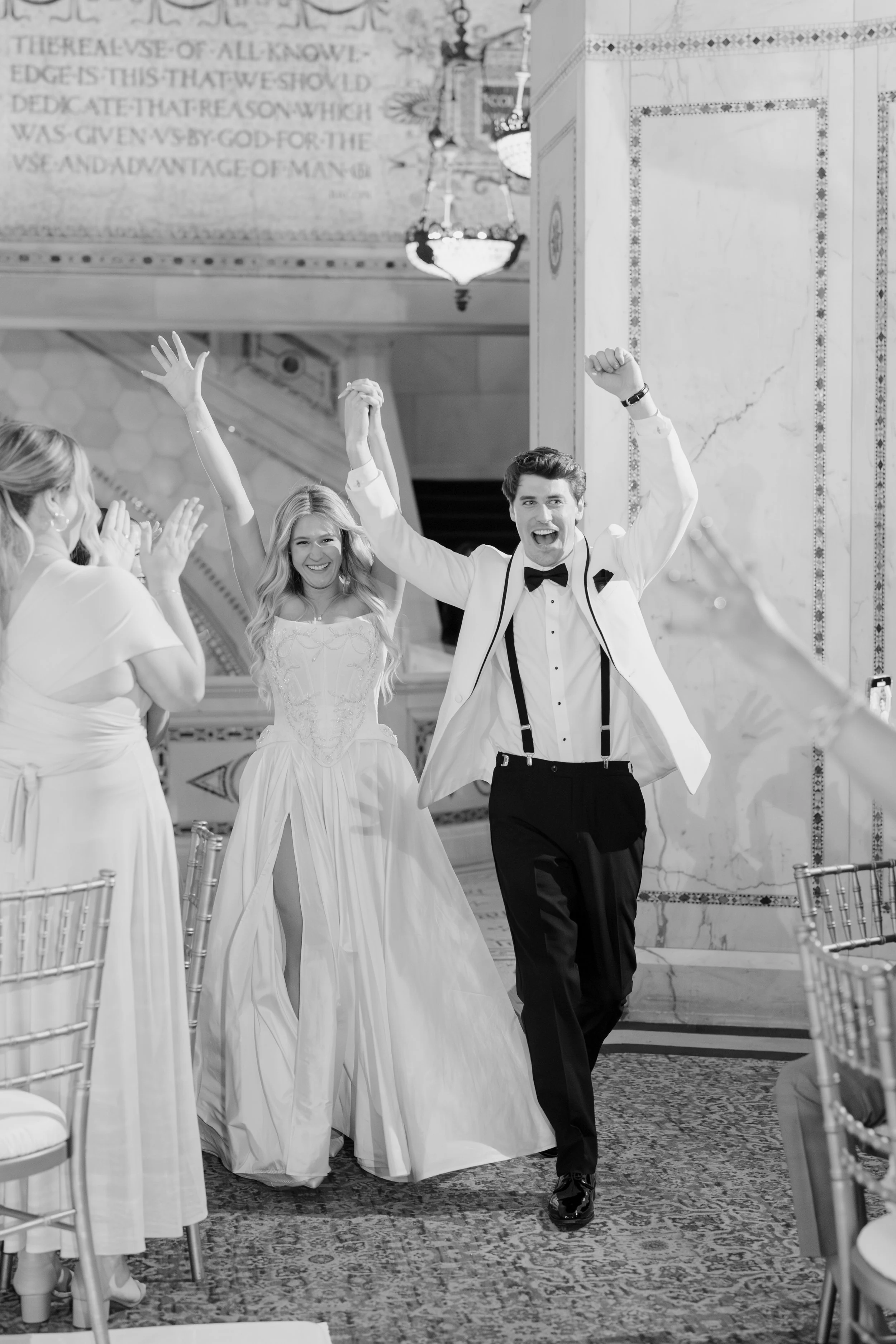 A man in a tuxedo with suspenders and a bowtie is celebrating with arms raised. A woman in a wedding dress is smiling nearby, and other women in dresses clapping and cheering. The scene is inside a decorated hall with chandeliers and ornate walls.