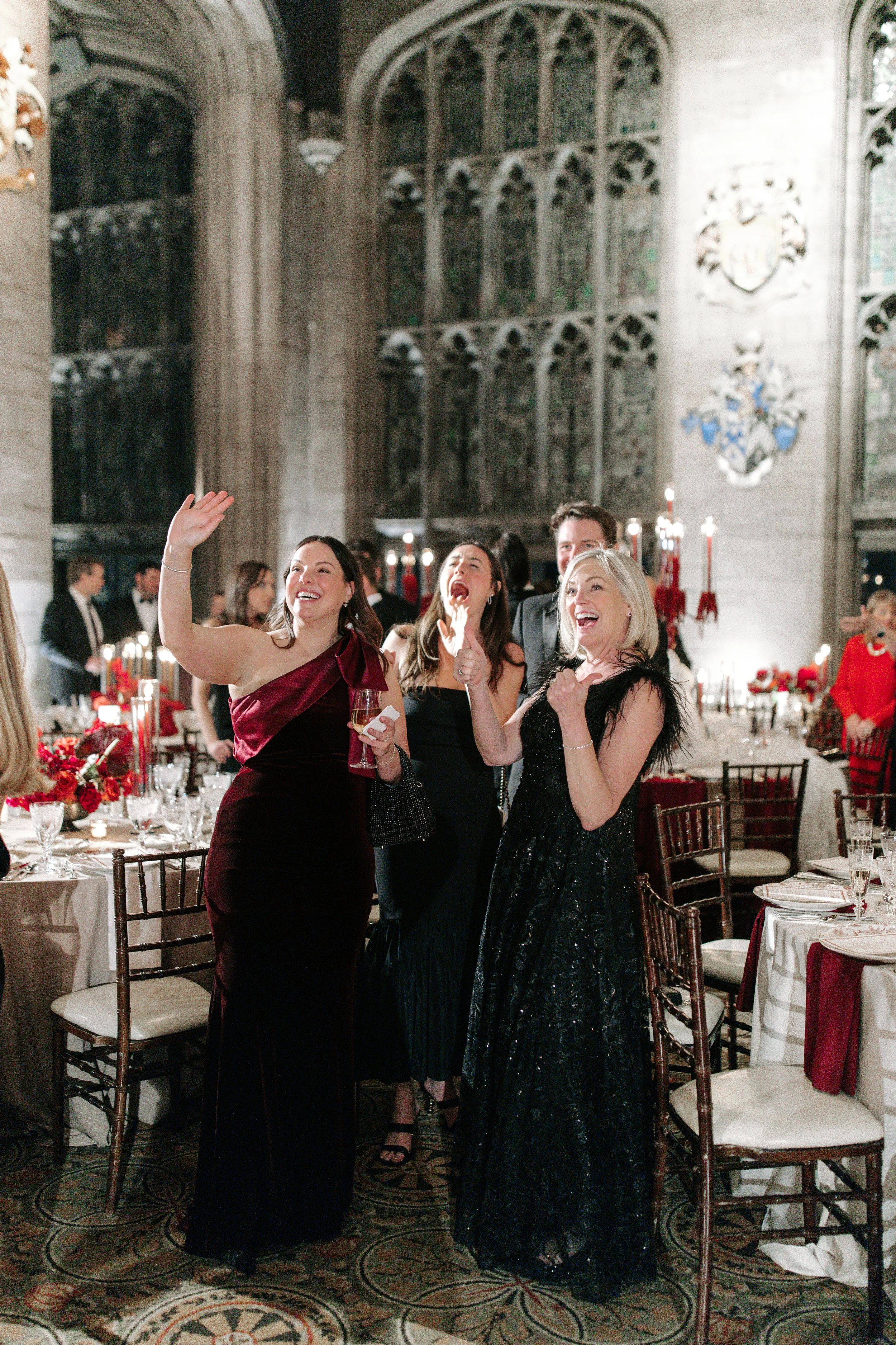 Group of people celebrating at a formal event in a grand hall with high ceilings, stained glass windows, and ornate decor. The women in the foreground are smiling and laughing, one in a burgundy gown, another in a black dress with feathers, and a wom