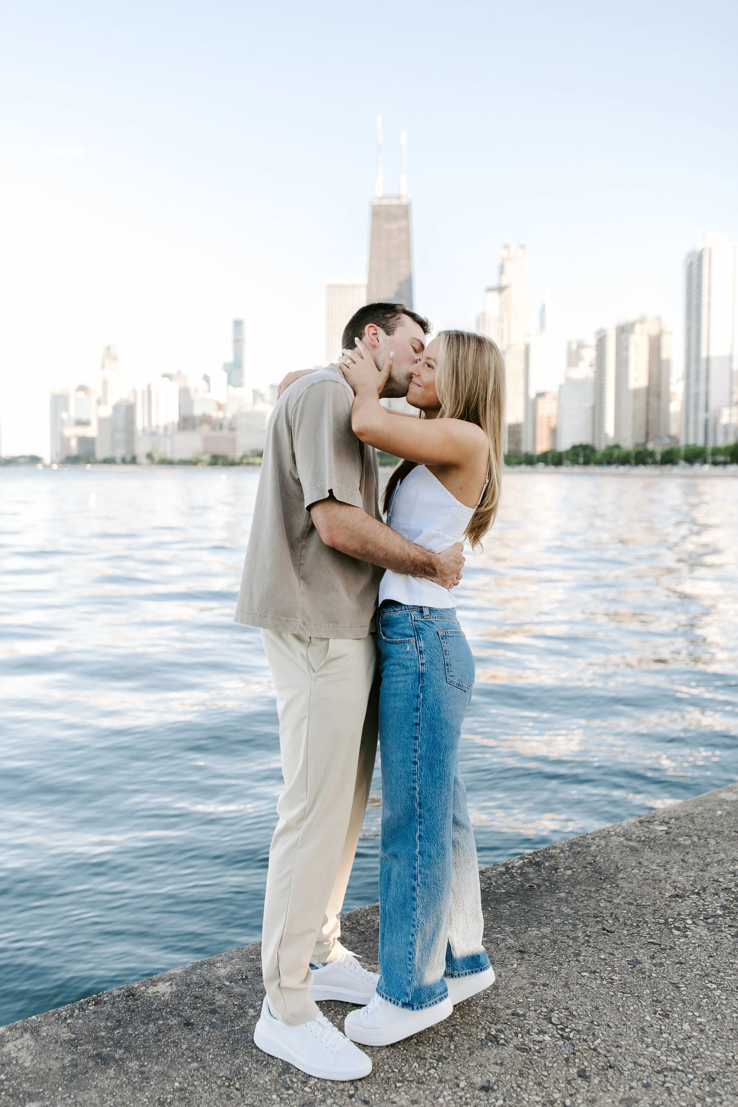 A couple hugging and kissing by a waterfront with a city skyline in the background.