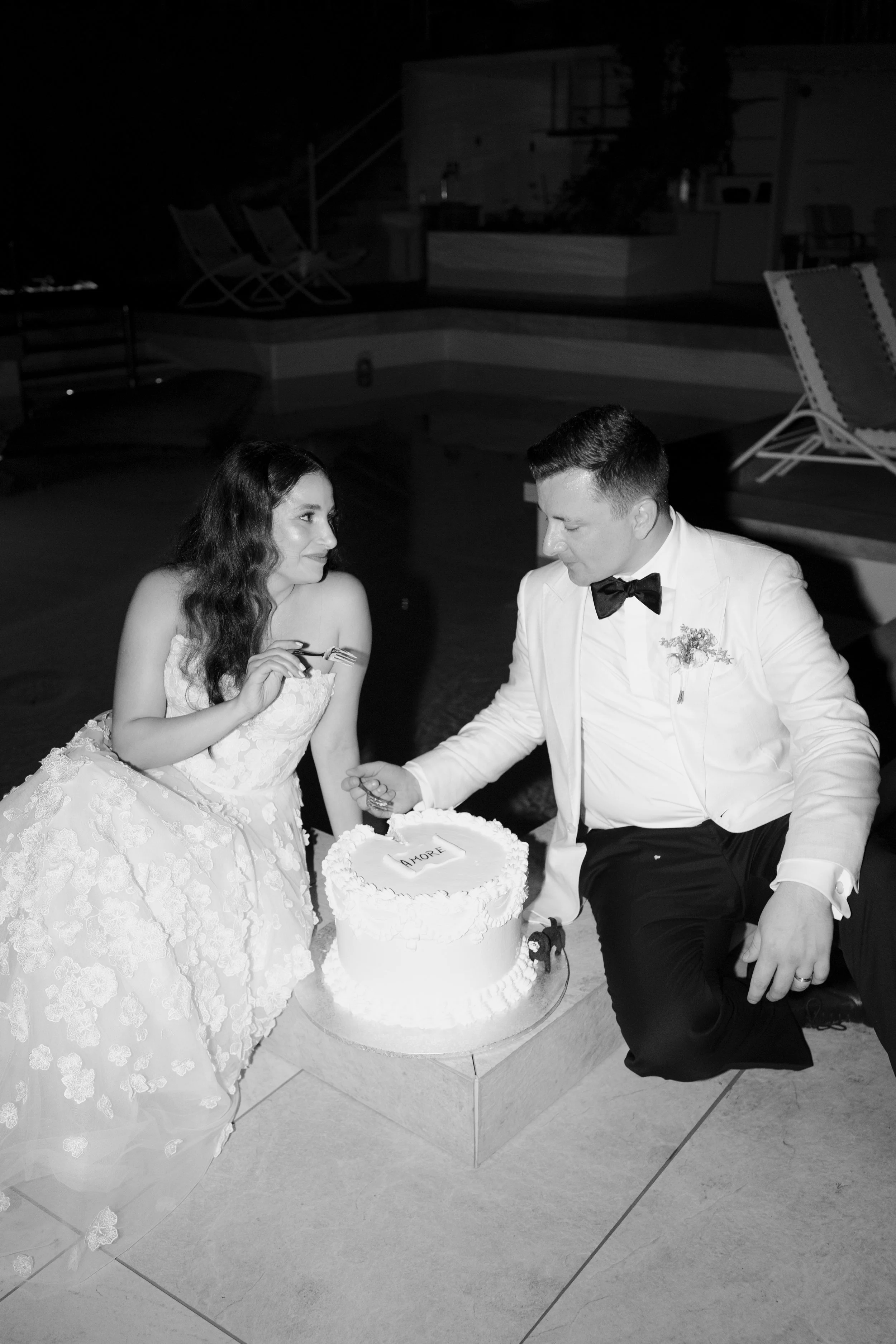 A bride and groom sit by a wedding cake, holding hands, with the bride holding a fork. The bride wears a lace wedding dress and looks at the groom. The groom is dressed in a tuxedo with a bow tie. The wedding cake has the word 'AMORE' written on it a