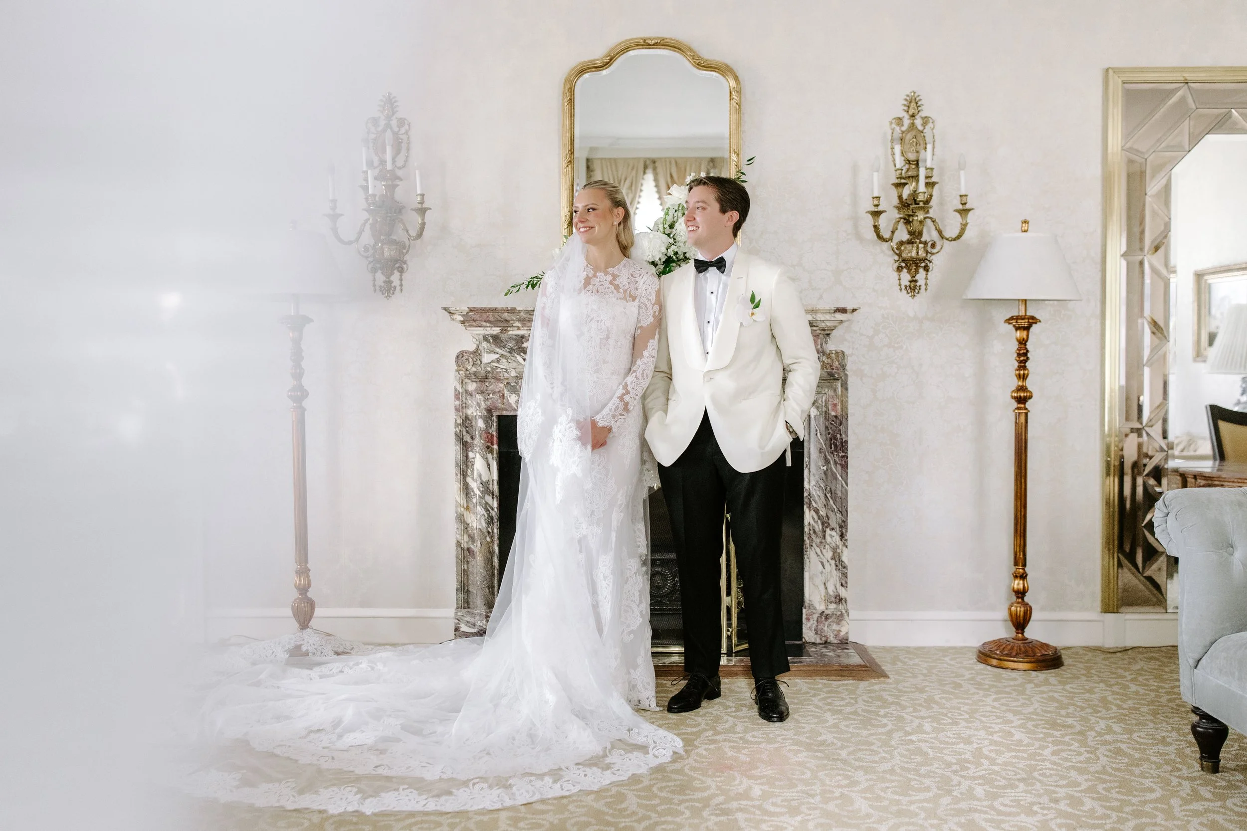 A bride in a lace wedding dress and a groom in a white tuxedo with a black bow tie standing together in a decorated room with a marble fireplace, mirrors, and gold wall sconces.