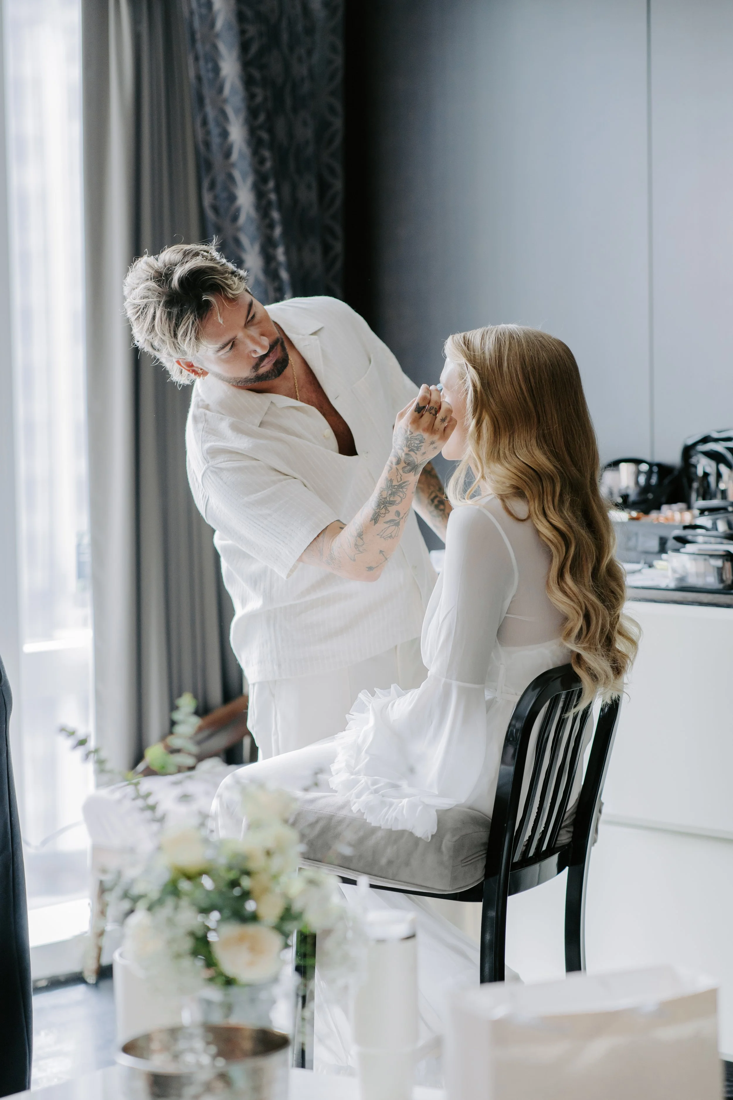 A makeup artist applying makeup to a young woman with long, wavy hair, sitting in a decorated room with natural light streaming in through the window.