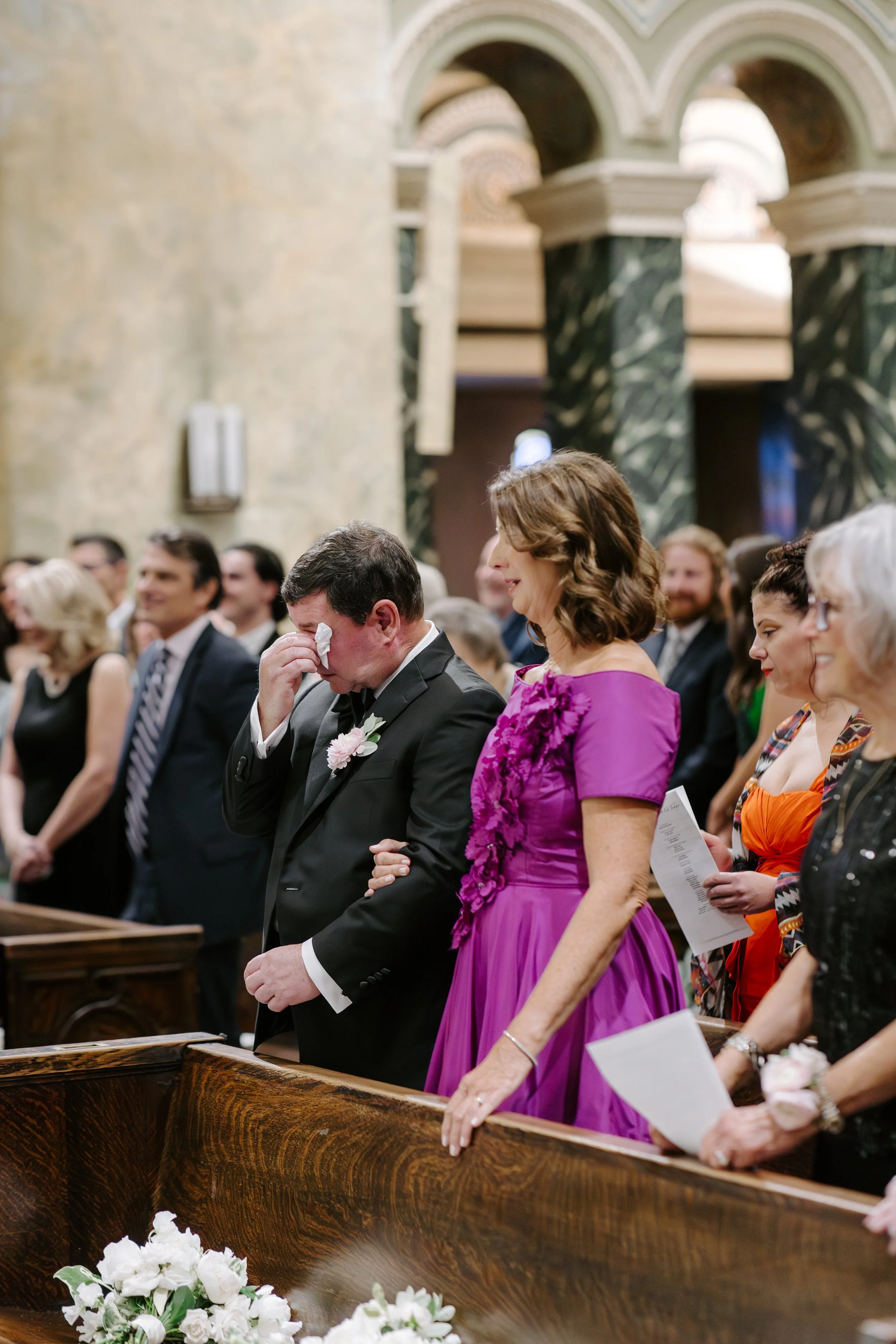 A group of people at a formal event, likely a wedding, inside a church. They are standing with heads bowed in prayer or reflection, and some are holding papers. The setting features ornate architectural details and floral arrangements.