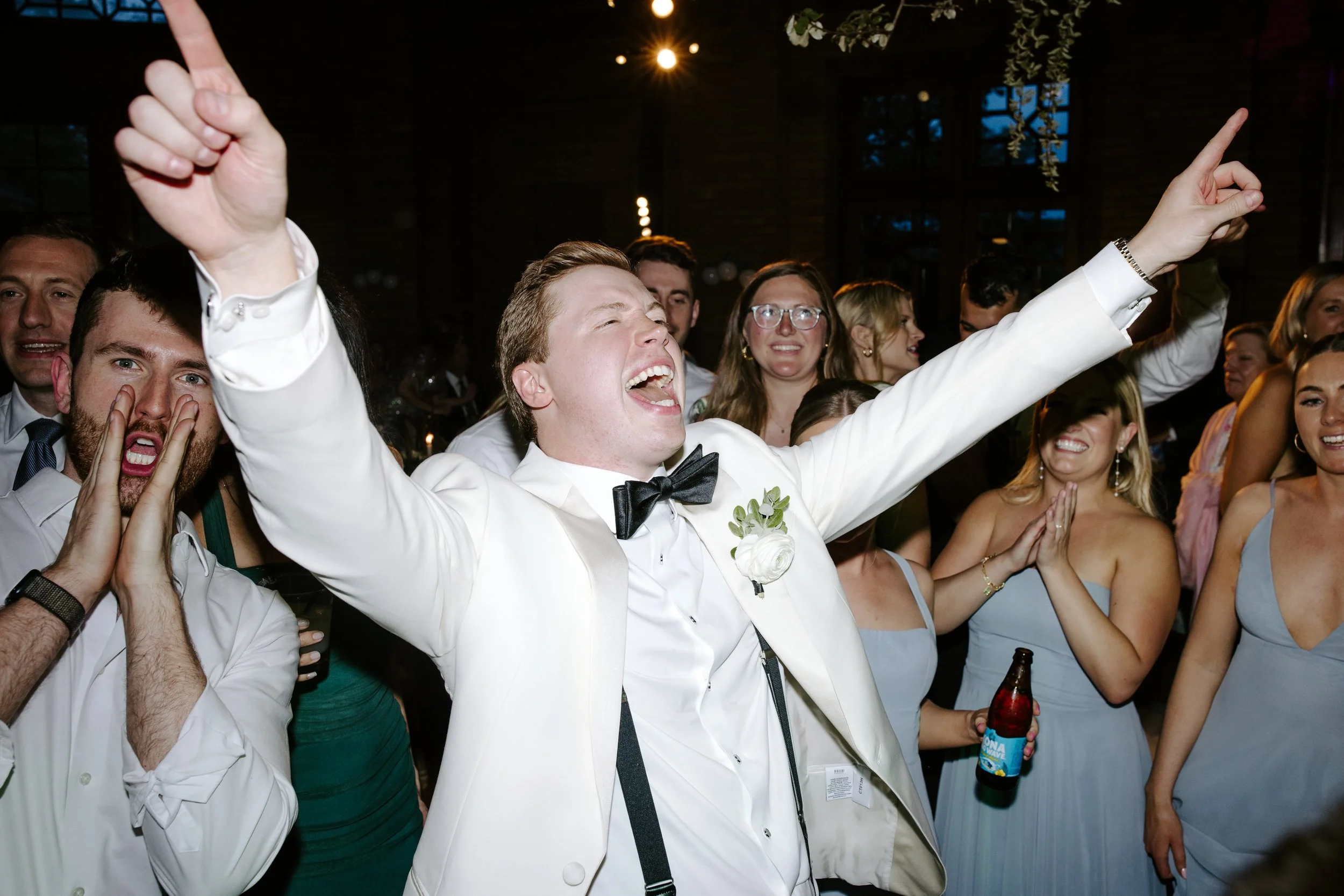 People at a celebration, smiling and dancing, with a man in a white suit and black bow tie at the center, raising his arms happily.