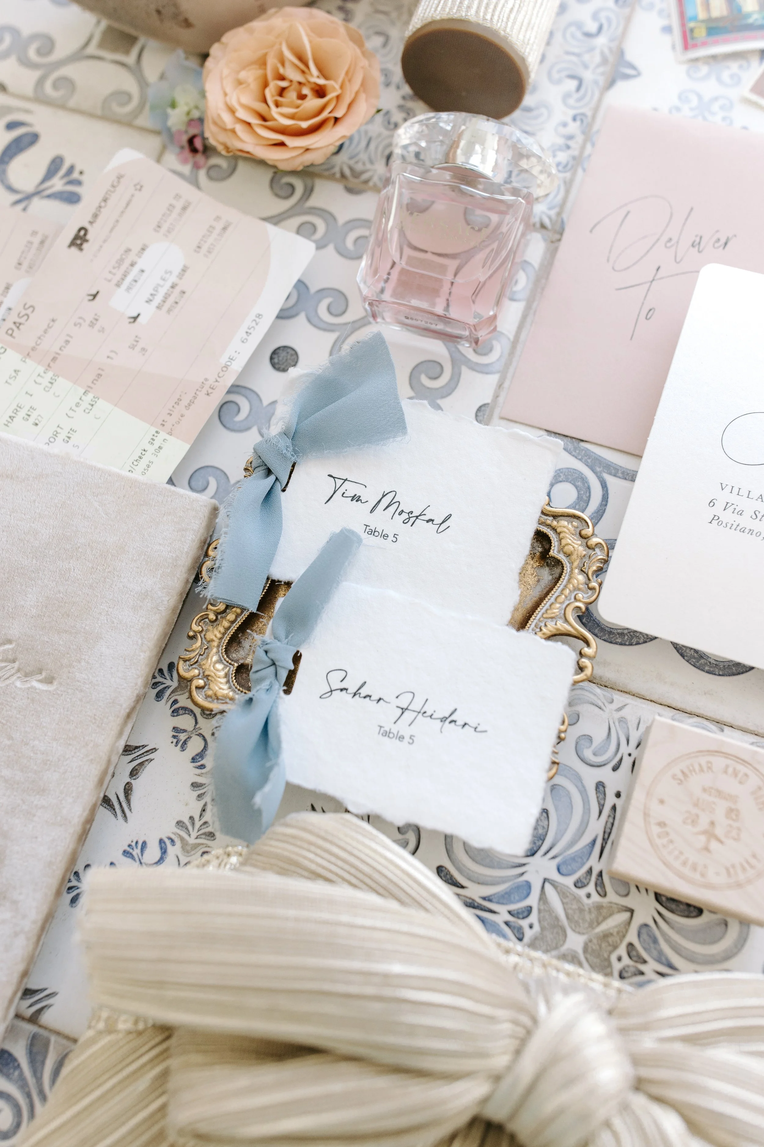 Wedding or event table setting with a small pink flower, perfume bottle, invitation cards labeled 'Sahar Heidari' and 'Tim Mosfal', and a train ticket, all arranged on a decorative silver surface.
