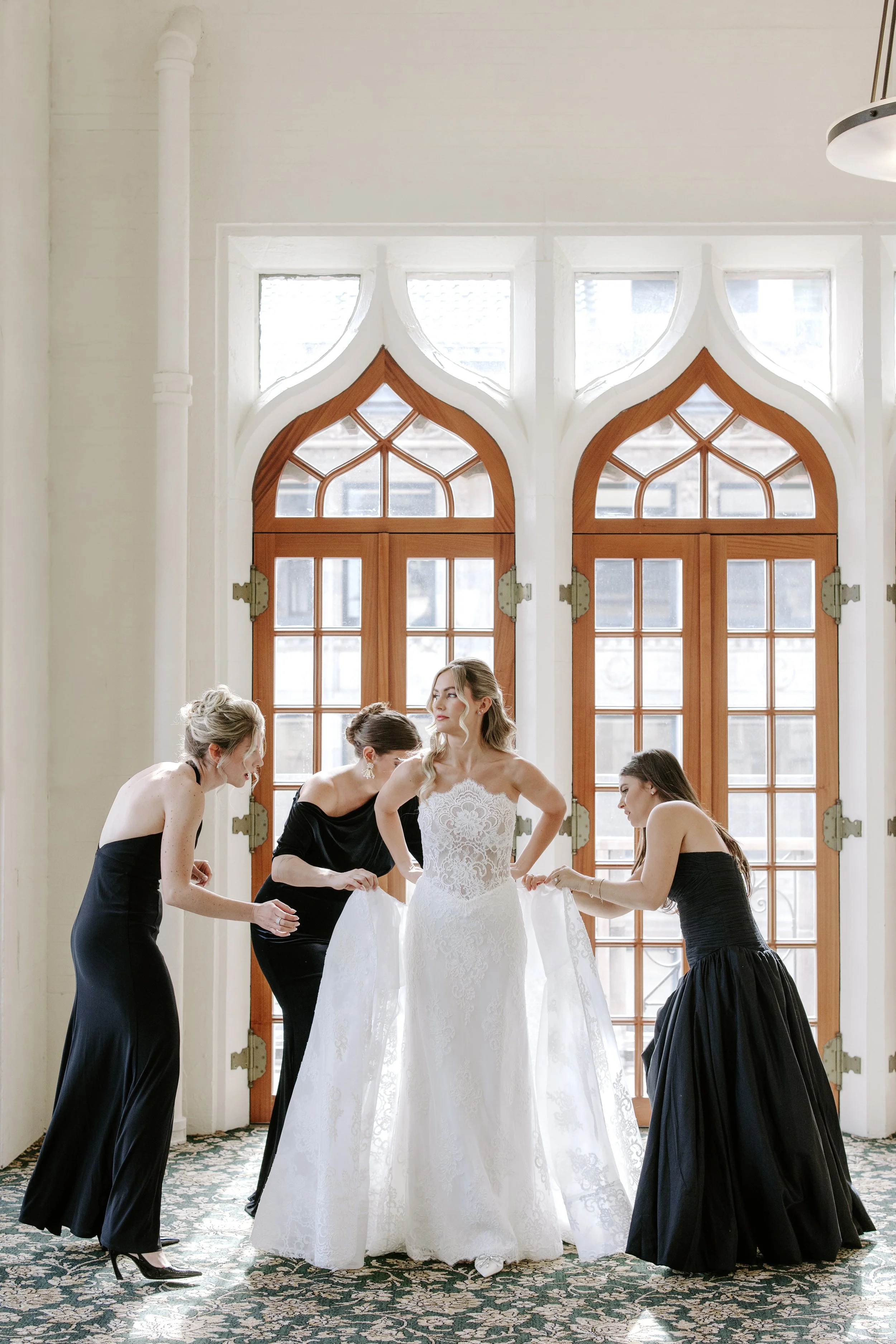 A bride is surrounded by five women in black dresses inside a bright room with large wooden doors and arched windows.