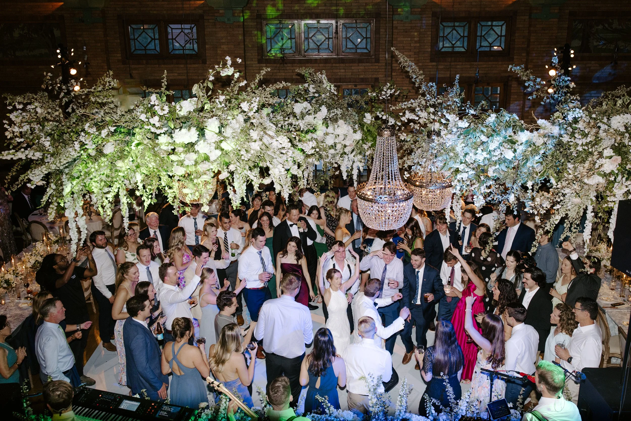 A large group of people dancing and celebrating at an indoor wedding reception with elaborate floral decorations and chandeliers.