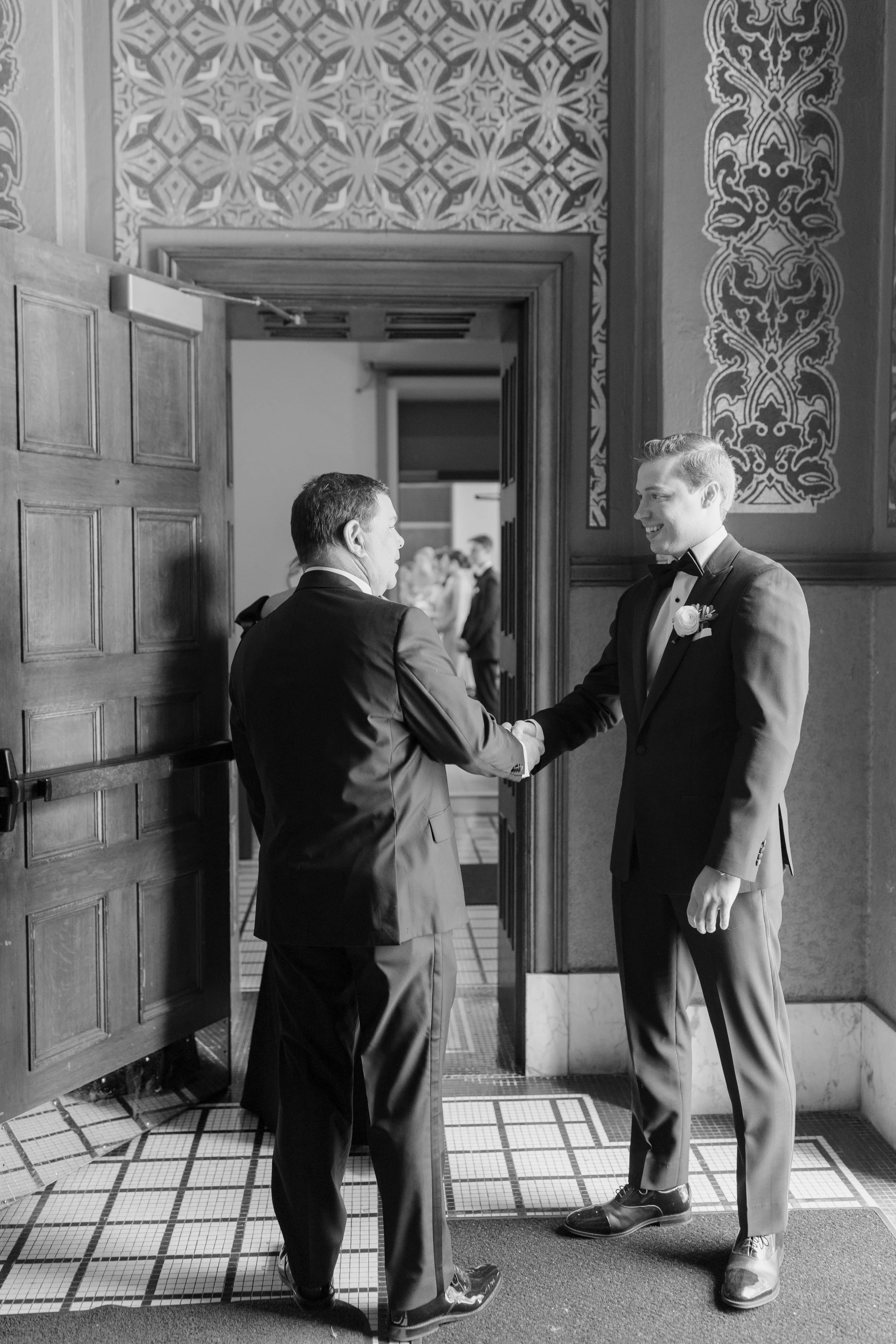 Two men in tuxedos shaking hands in a formal setting. One older man with dark hair facing away from camera, and a younger man with light hair smiling, wearing a boutonniere. The room features decorative wallpaper, wooden door, and a tiled floor.