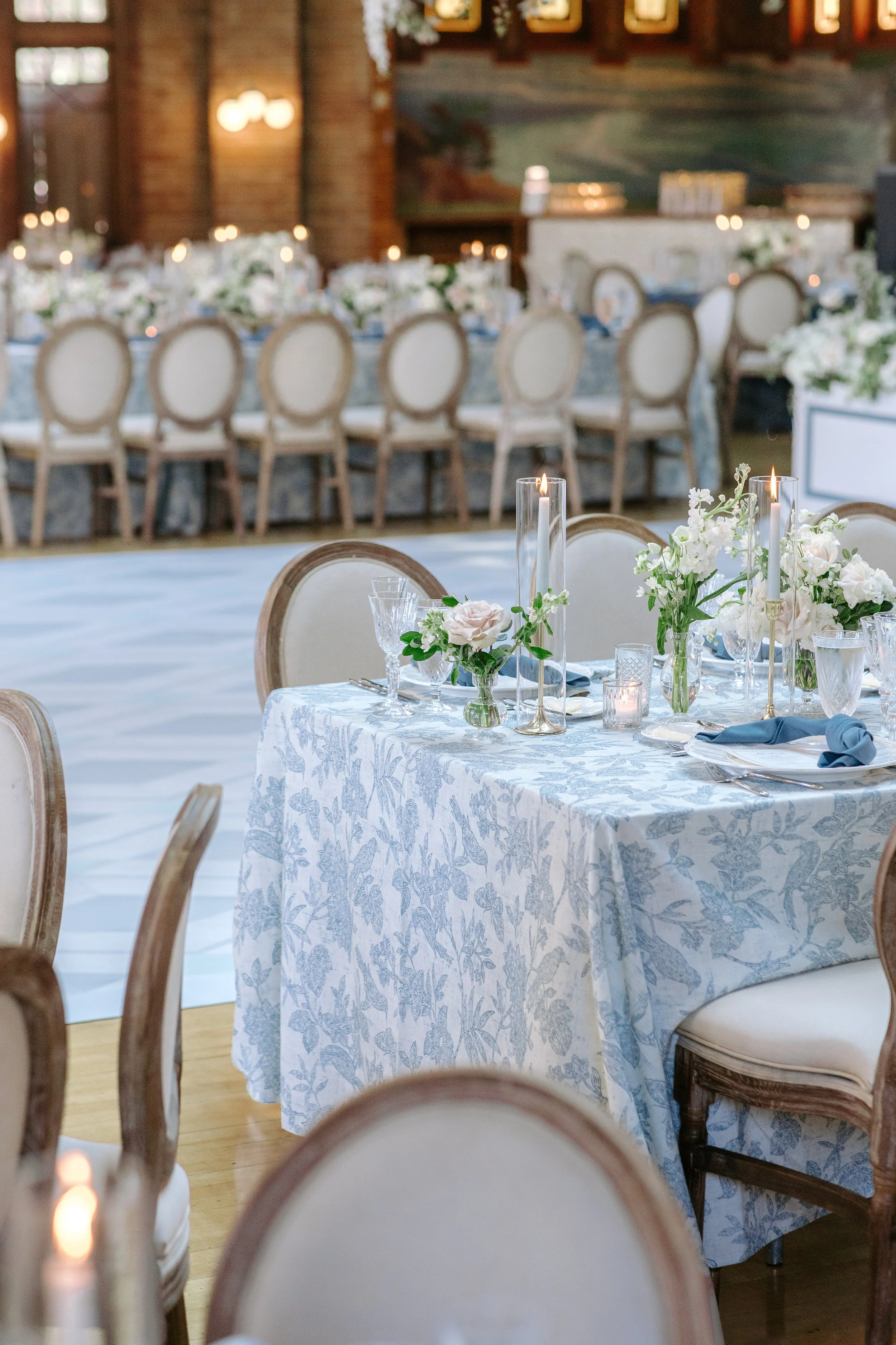 Elegant wedding reception with a round table covered in blue and white floral tablecloth, adorned with white and blush pink flowers, tall taper candles, and glassware. In the background, there is a long banquet table with similar floral centerpieces 