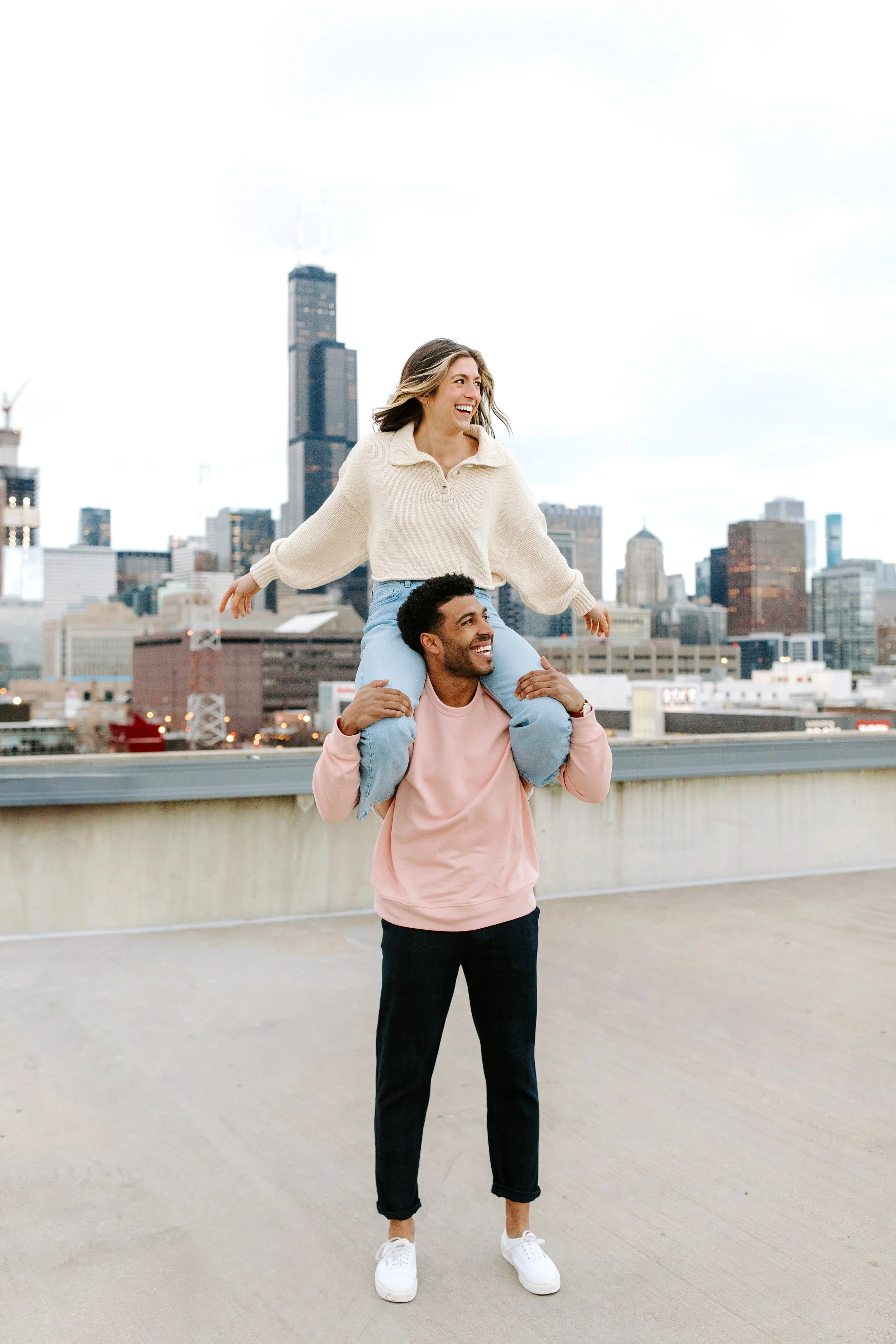 A man with a beard and dark hair wearing a pink sweatshirt and white sneakers is carrying a woman with long hair, wearing a cream sweater and light blue jeans, on his shoulders on a rooftop with a city skyline in the background.