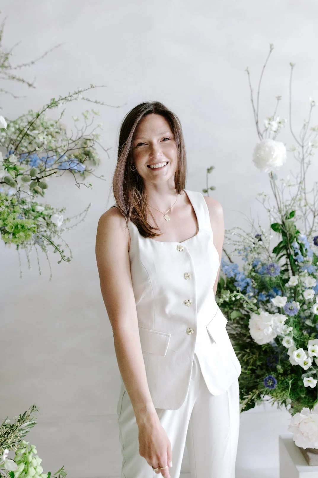 A woman with long brown hair wearing a white sleeveless blazer and matching pants, smiling, standing among white, blue, and green floral arrangements in a bright setting.