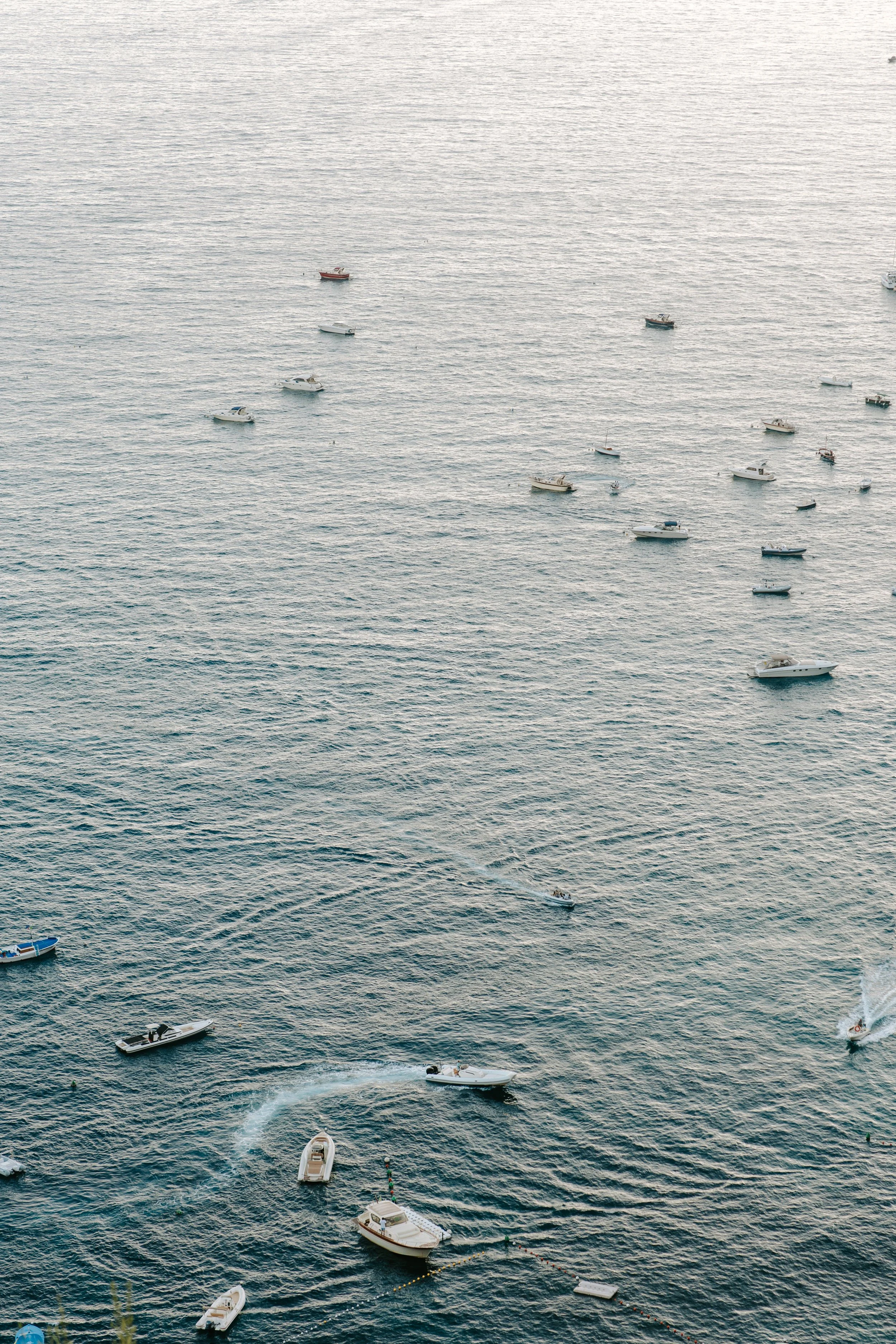Multiple boats and yachts floating in calm ocean water, with some boats creating ripples and wake in the water.