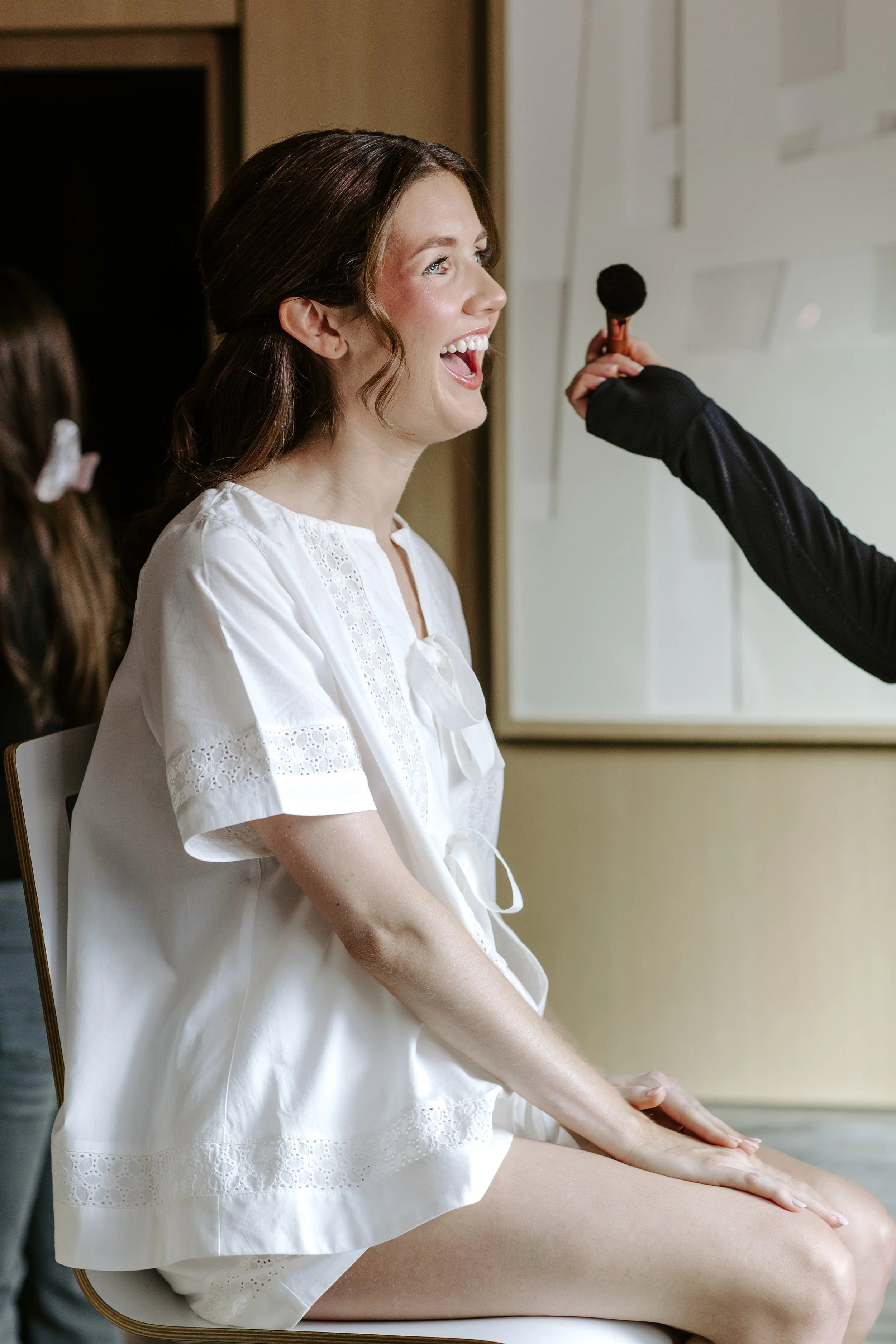 A woman in a white dress sitting on a massage table, smiling and having her leg massaged by a person in black clothing with a black massage tool in hand.