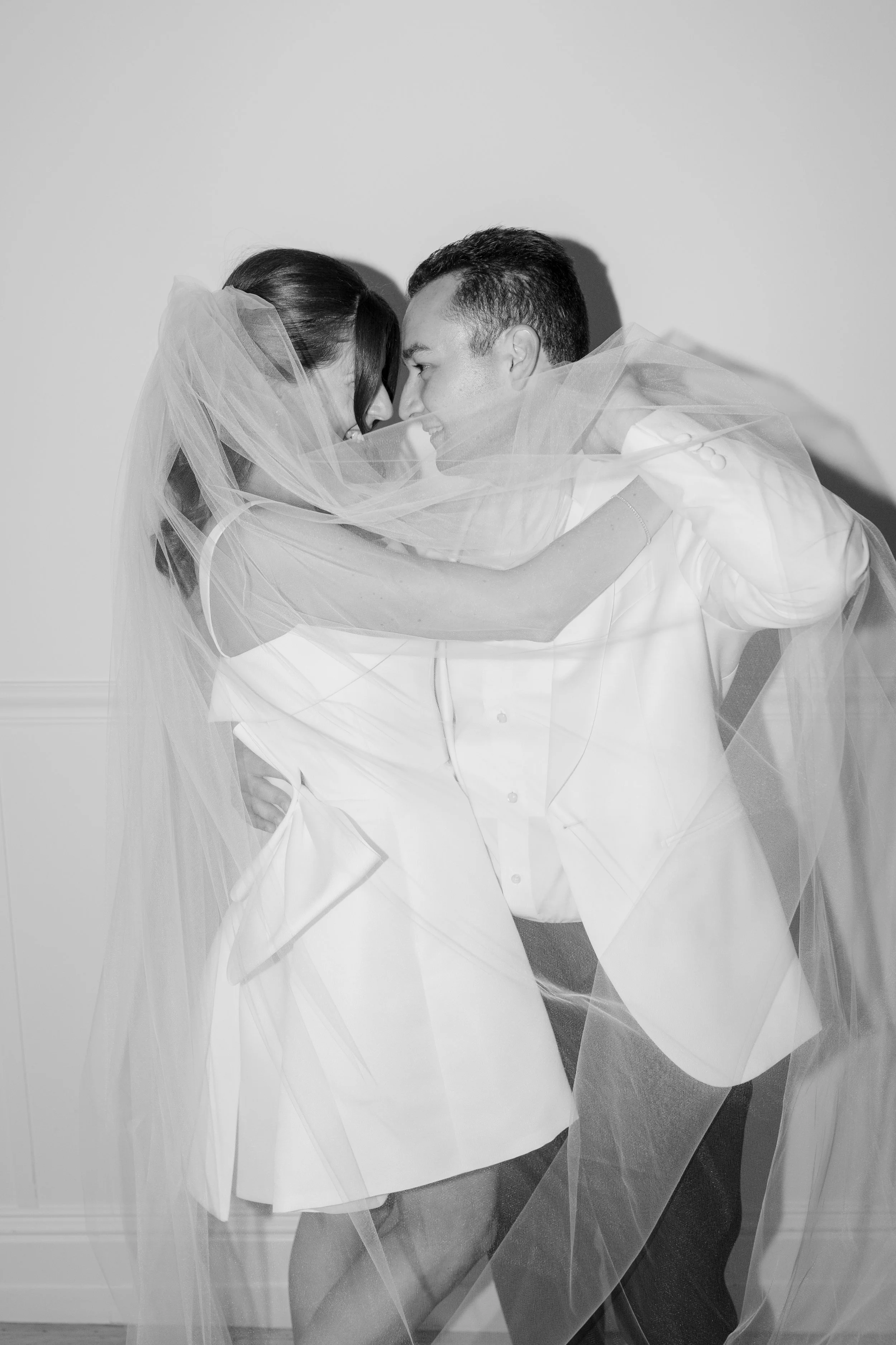 A black-and-white photo of a couple with light skin and dark hair, wrapped in a sheer veil, looking into each other's eyes, smiling, indoors against a plain wall.