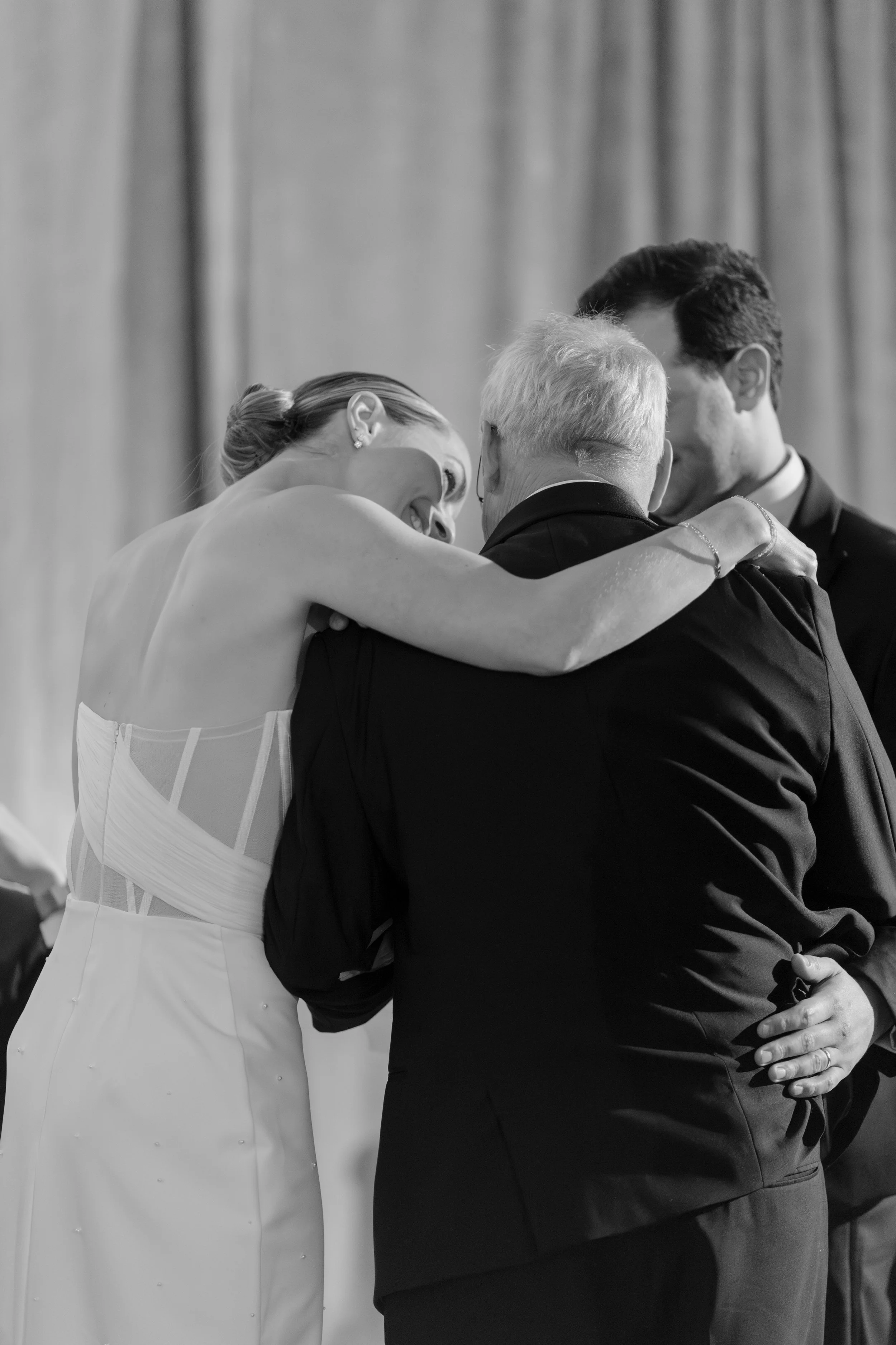 A black and white photograph of a bride and two male figures, possibly the officiant and groom, sharing a moment during a wedding ceremony. The bride is smiling and leaning in towards the man on the right, with her arms around his shoulders. The man 